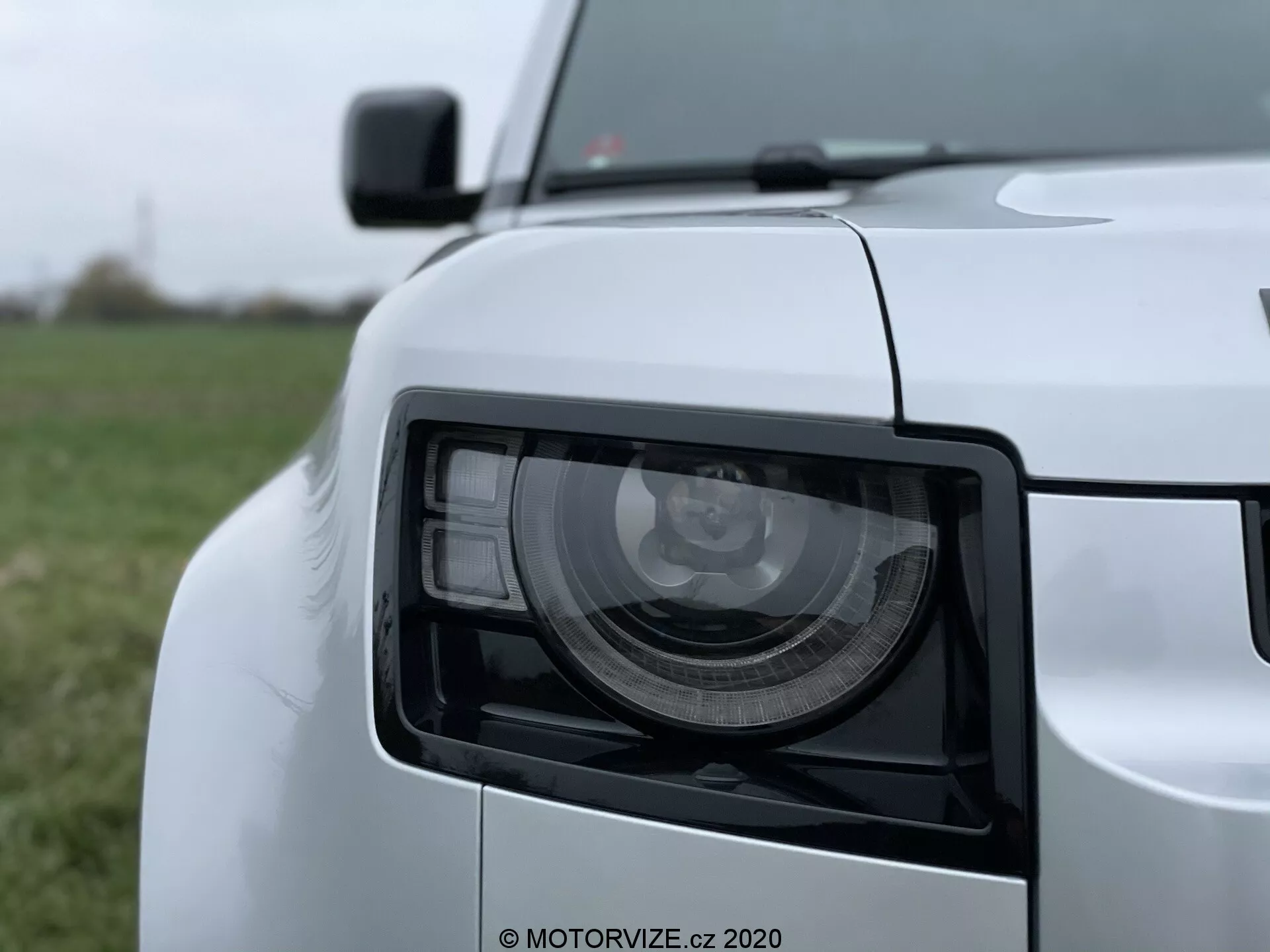 Vista de close-up do conjunto do farol dianteiro esquerdo do Land Rover Defender 2020 com design angular moderno, tirada em um ângulo leve com perspectiva de baixa a média altura. A imagem mostra os módulos de luz LED/HID detalhados, acabamento em pintura branca brilhante e parte da grade dianteira desfocada, borda do capô, seção do para-choque dianteiro e espelho lateral.
