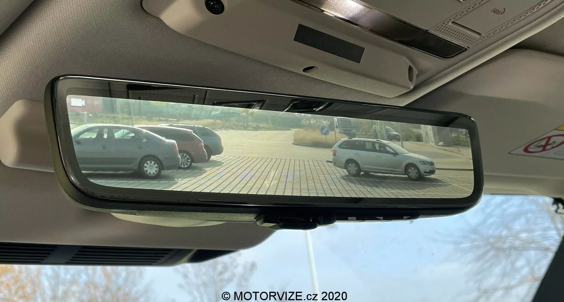 Vista do assento do motorista de um Land Rover Defender 2020, focada no espelho retrovisor interno retangular com escurecimento automático que reflete dois hatchbacks prateados estacionados perpendicularmente em um estacionamento pavimentado com pedras bege em um dia ensolarado.