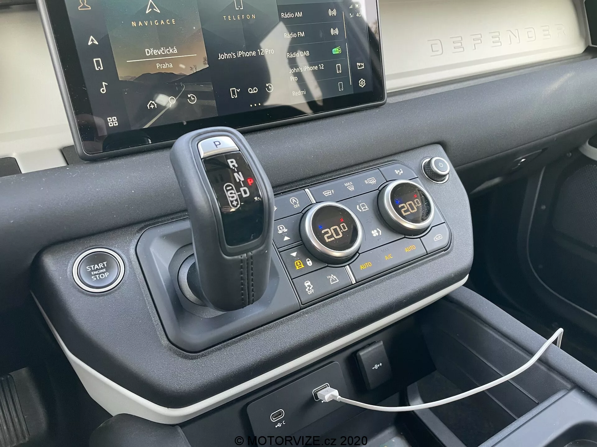 Close-up da consola central do Land Rover Defender 2020 destacando a alavanca de mudanças, o sistema de resposta ao terreno para várias condições de condução, o botão de Start/Stop do motor, os controlos de climatização de duas zonas, a porta USB para conectividade de dispositivos, o sistema de infotainment com ecrã tátil parcialmente visível e os materiais premium utilizados no design interior, visto a partir da perspetiva de um assento dianteiro num ângulo de 45 graus.
