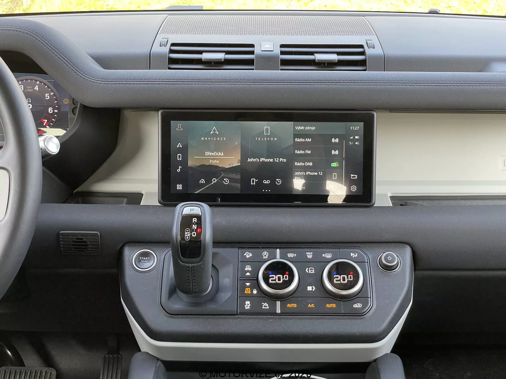 Vista do painel interior do Land Rover Defender 2020 mostrando o sistema de infotainment com tela sensível ao toque, saídas de ar duplas, seletor de marchas moderno, painel de controle climático de duas zonas, vislumbre do volante e uma combinação de plásticos rígidos e materiais macios ao toque com um contraste de cores no painel.
