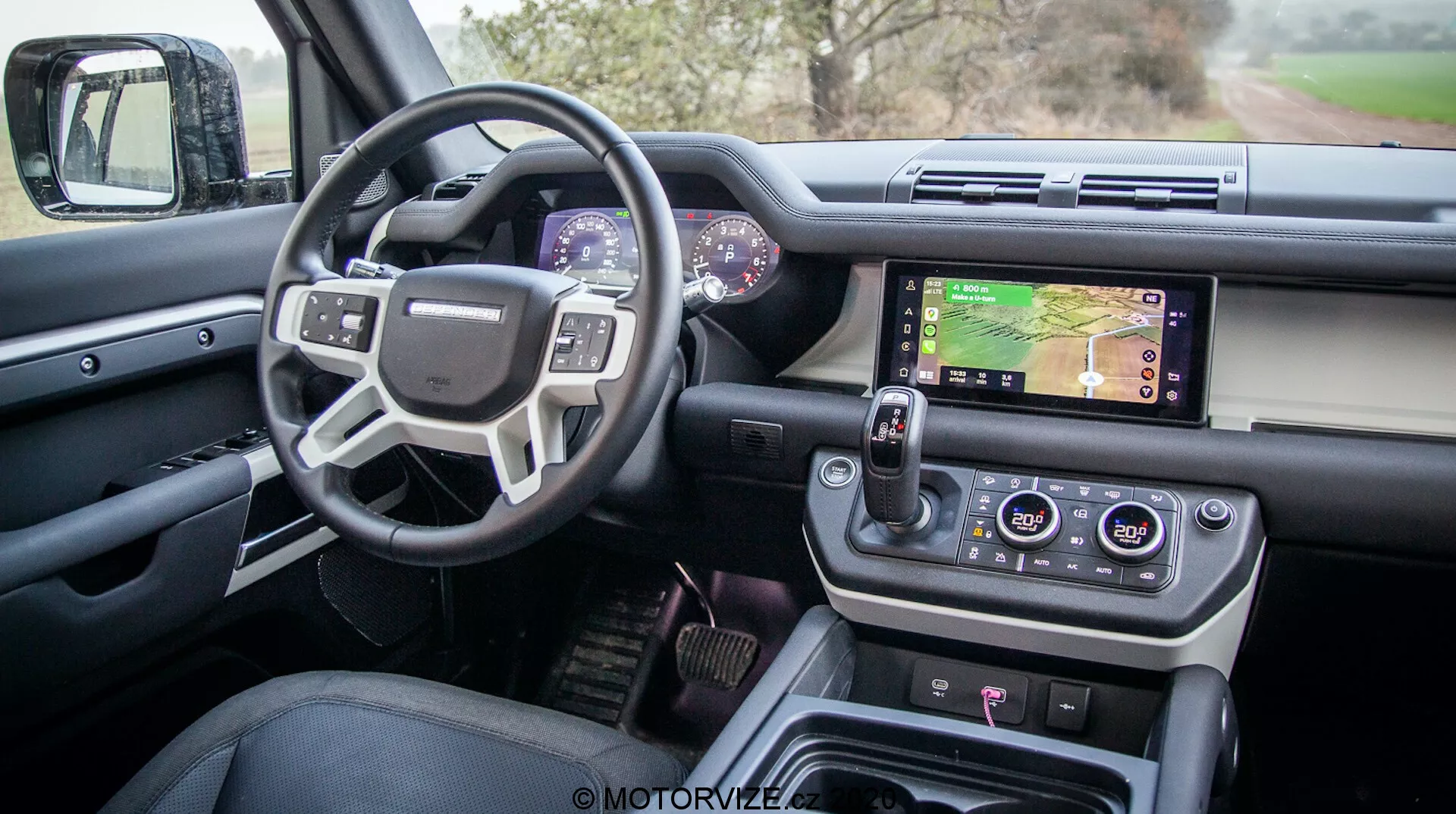 Vista interna de um moderno Land Rover Defender 2020 pela porta do motorista, mostrando o design sofisticado e funcionalidades avançadas, incluindo uma porta do motorista aberta com controles para vidros e espelhos, volante multifuncional com botões de controle, painel de instrumentos digital, sistema de infotainment central com tela sensível ao toque mostrando um mapa, controles de climatização, seletor de marchas rotativo moderno e botões de controle de terrenos, tudo em um acabamento de luxo de alta qualidade.