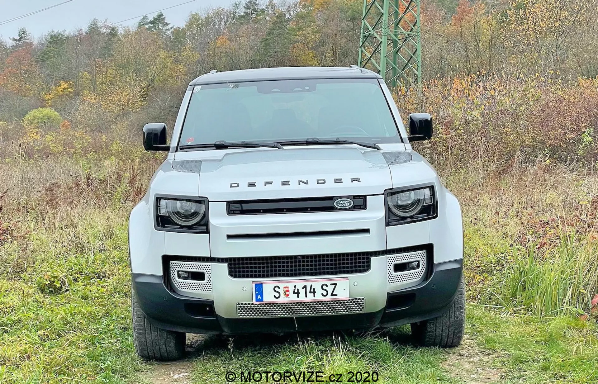 Vista frontal de um Land Rover Defender 2020 branco 4x4, com uma grelha retangular proeminente ladeada por faróis LED redondos com luzes diurnas quadradas integradas, para-choques dianteiro robusto com aberturas retangulares, capô plano com a inscrição 'LAND ROVER' e a marca 'DEFENDER' na borda frontal, para-brisa plano, placa de licença europeia montada centralmente e sensores de estacionamento, capturada estacionada em um ambiente natural.