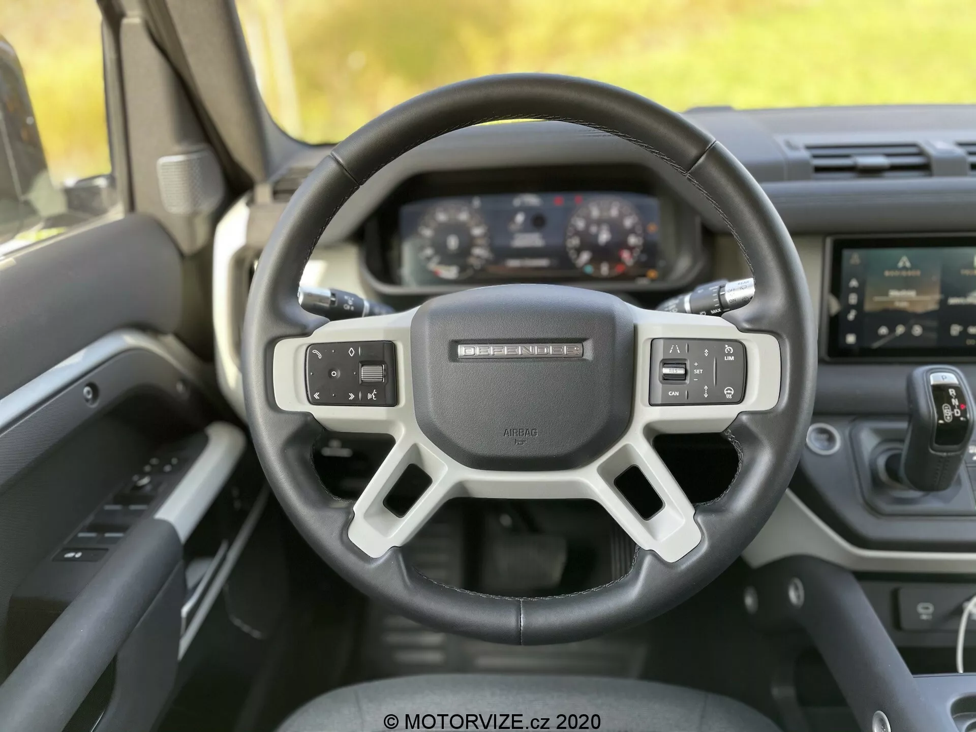 Vista interna de um Land-Rover Defender 2020 a partir da perspectiva do motorista, destacando o volante moderno de três raios com controles integrados para áudio, comandos de voz e configurações de controle de cruzeiro. No painel de instrumentos atrás, vemos displays digitais para velocímetro, tacômetro e uma tela de múltiplas informações. A imagem também captura o console central com um sistema de infotainment touchscreen e botões de controle de clima abaixo. O ângulo da foto é ligeiramente inclinado para cima, destacando o design ergonômico, a tecnologia avançada e os materiais premium do veículo estacionário.