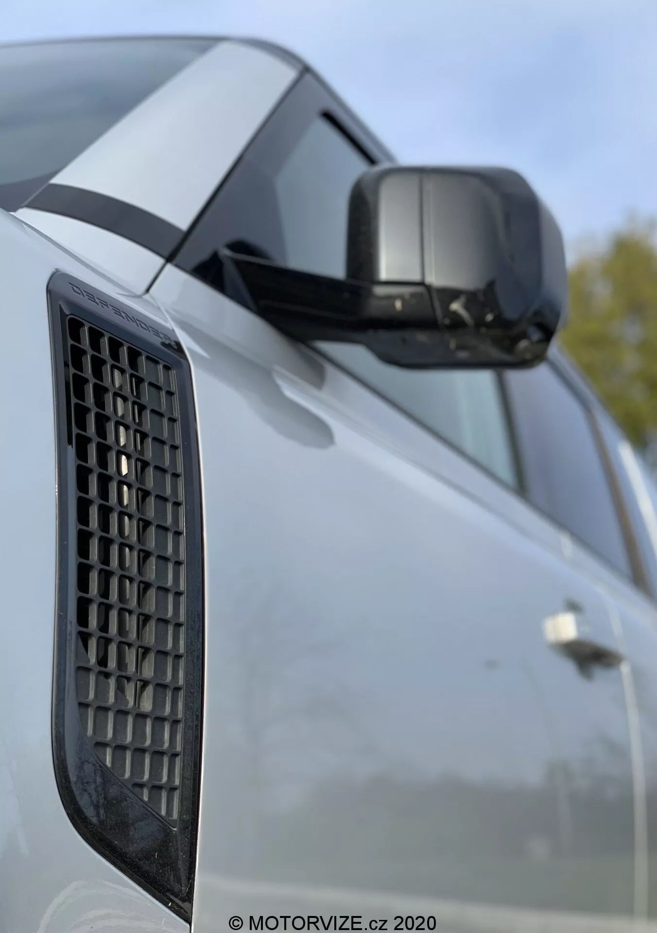 Close-up do Land Rover Defender 2020 destacando o para-lama lateral com uma grade, o espelho lateral com indicador de direção integrado e uma porção da porta com linhas de caráter marcantes; a imagem foca nos elementos de design do veículo com um fundo desfocado sugerindo um ambiente externo.