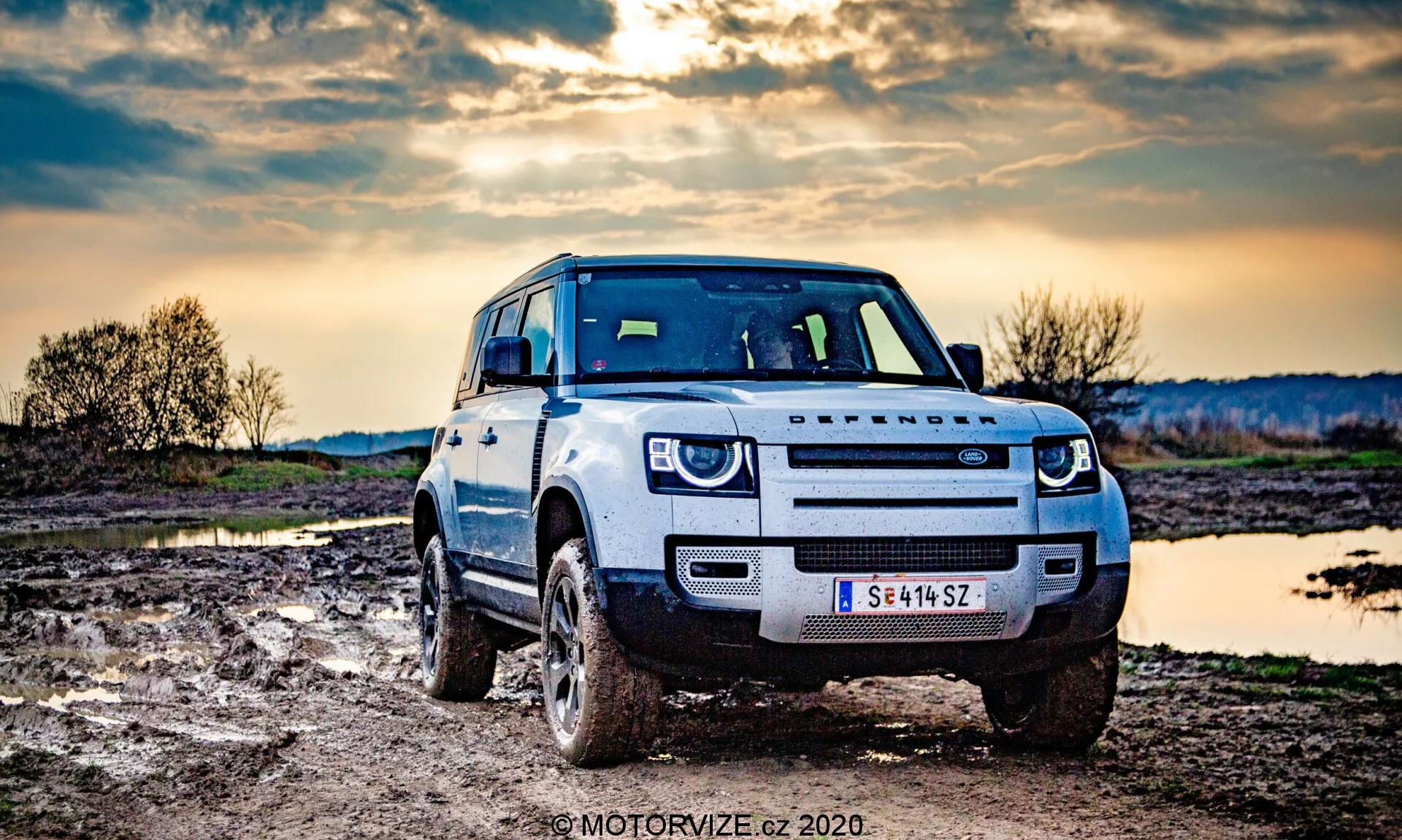 Fotografia de um SUV Land Rover Defender tirada de um ângulo frontal, ligeiramente descentralizado, destacando a parte frontal do veículo e sua capacidade off-road. O veículo é de cor clara, com sujeira nos pneus e nos painéis inferiores, indicando que foi dirigido fora de estrada. O carro tem um snorkel para atravessar águas profundas e faróis de LED. O ambiente rural apresenta terreno lamacento e um corpo de água parada, com um céu dramático ao entardecer ao fundo.