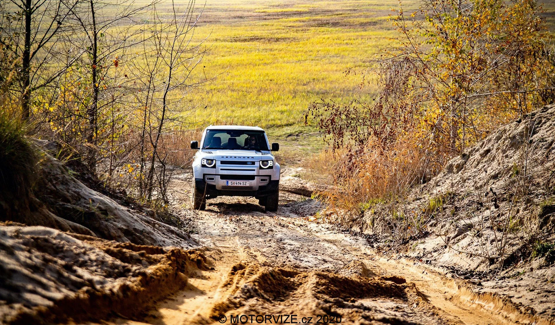 Um SUV Land Rover Defender 2020 é visto de frente, dirigindo off-road, posicionado centralmente contra um pano de fundo de terreno irregular e ambiente natural. A parte frontal visível do carro mostra a articulação avançada da suspensão, faróis de LED e uma grade característica com ripas horizontais. Capturando o SUV em um ambiente rural com tonalidades de outono e luz natural, a perspectiva realça a presença robusta e aventureira do veículo.