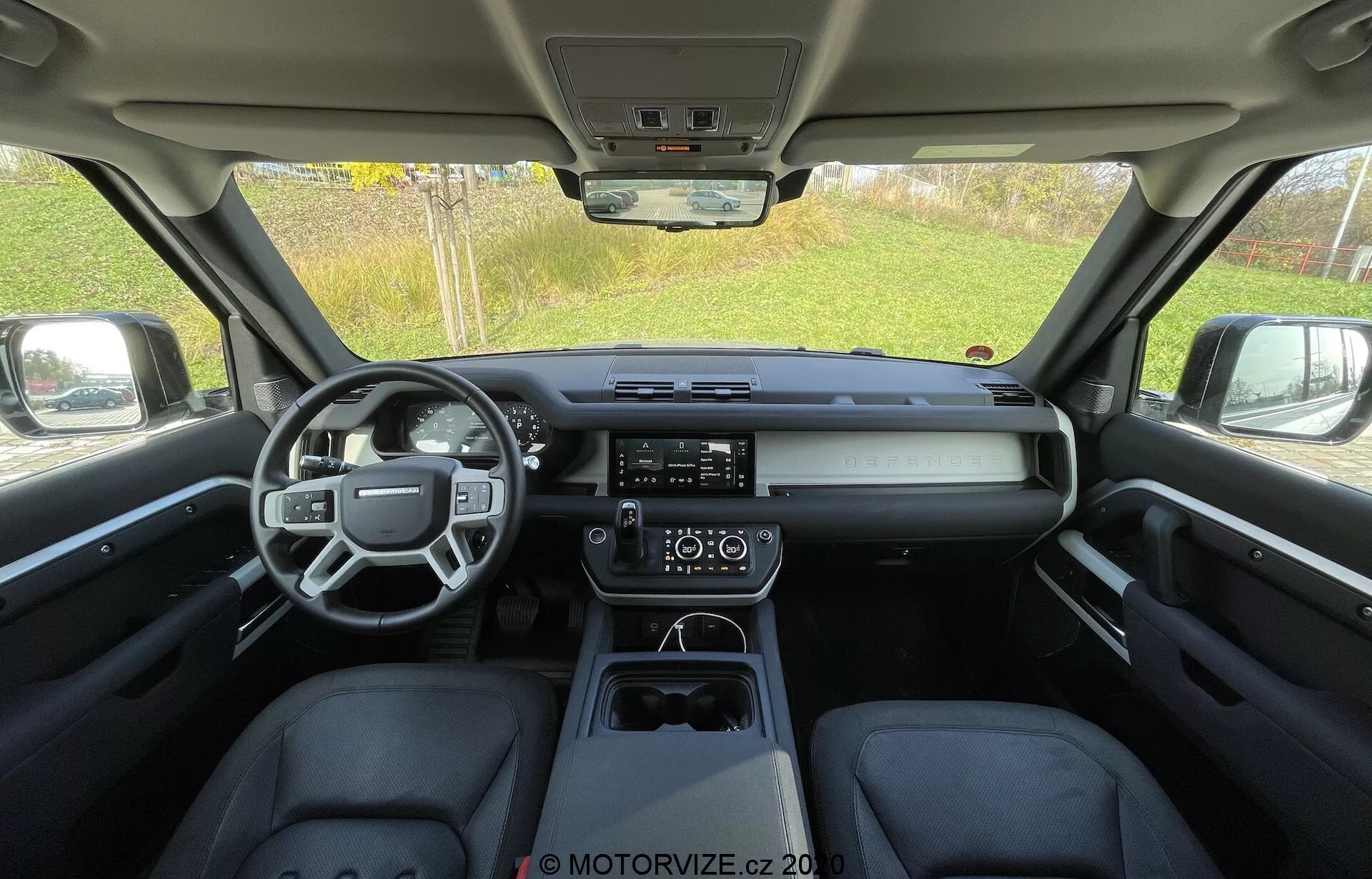 Vista interior de um Land Rover Defender 2020 a partir do banco traseiro, mostrando uma cabine elegante e moderna com um volante multifuncional, painel horizontal com um display digital central, sistema de infotainment com tela sensível ao toque proeminente, controle climático de duas zonas e um console central limpo com seletor de marcha eletrônico e porta-copos.
