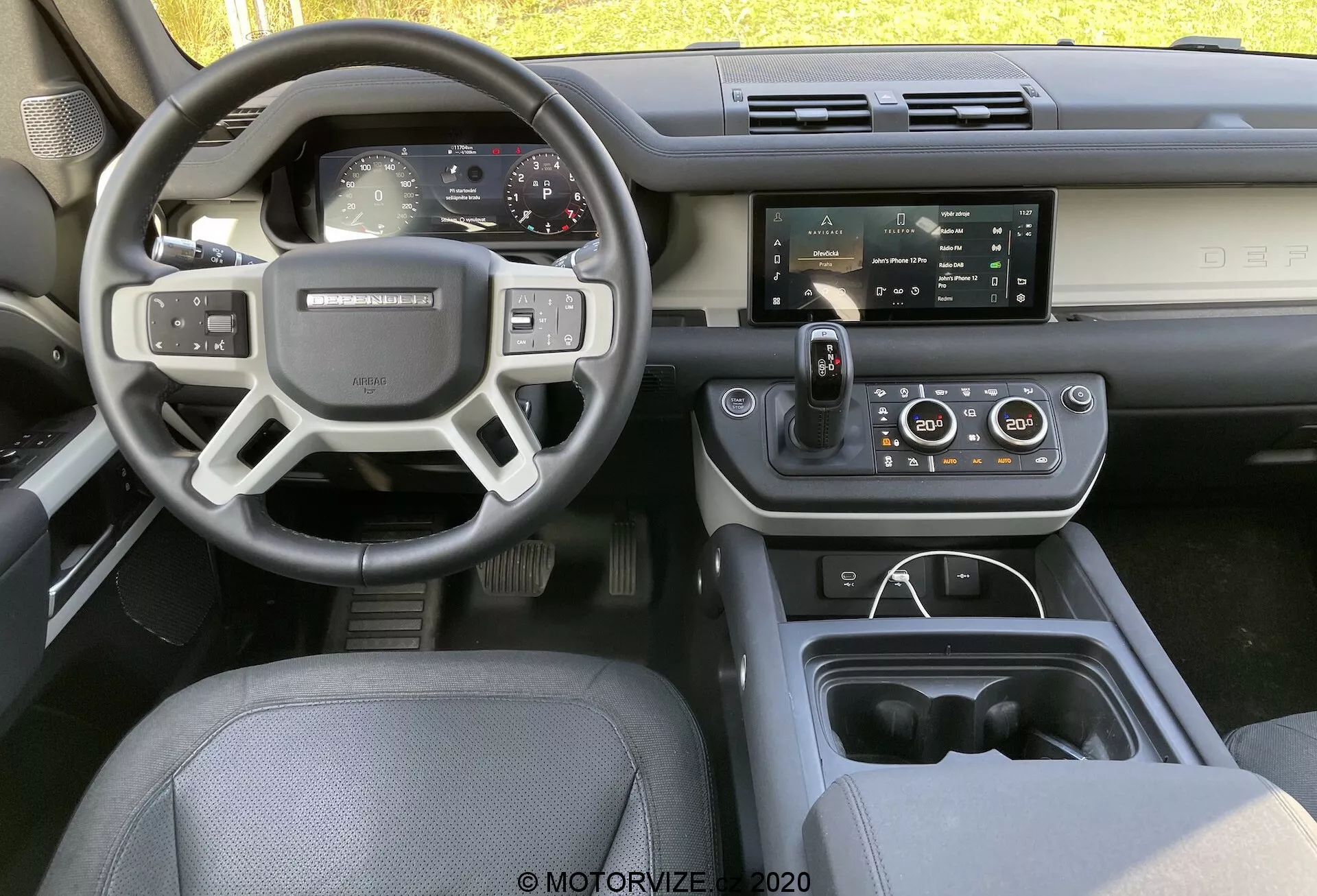 Imagem do interior de um Land Rover Defender 2020 a partir do assento traseiro central, mostrando o painel de instrumentos, volante com botões de controle, painel totalmente digital e tela central de infoentretenimento com tecnologias inteligentes e de conforto.