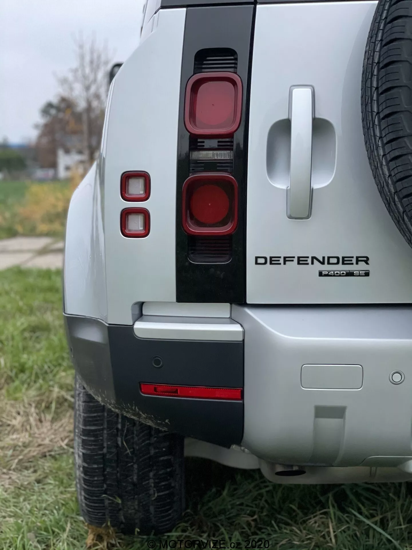 Close-up da seção traseira de um Land Rover Defender 2020 mostrando luzes traseiras modernas empilhadas verticalmente, emblemas proeminentes 'DEFENDER' e 'X-Dynamic SE', visão parcial do pneu sobressalente na porta traseira, para-choque de duas cores com um possível gancho de reboque e acabamento em prata, destacando o design premium e orientado para off-road do veículo.