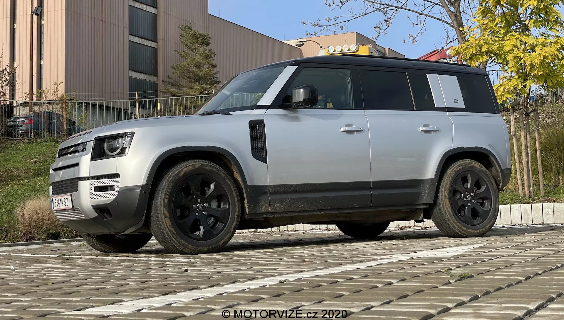 Fotografia de um SUV Land Rover Defender 2020 moderno tirada de um ângulo baixo para acentuar sua postura robusta e imponente. O veículo é mostrado em uma vista de três quartos da frente e do lado, destacando sua grade distinta, faróis de LED, para-choque com luzes de nevoeiro e sensores, rodas de liga leve pretas com design multirraiado e pneus de perfil baixo, espelhos laterais pretos com tecnologias adicionais potenciais, vidros escurecidos, linha de teto proeminente, maçanetas das portas embutidas, design forte dos arcos das rodas e cor prata clara ou cinza contrastando com os elementos de acabamento preto. O SUV está estacionado em um pavimento de paralelepípedos, com um edifício e folhagem ao fundo sob iluminação diurna.