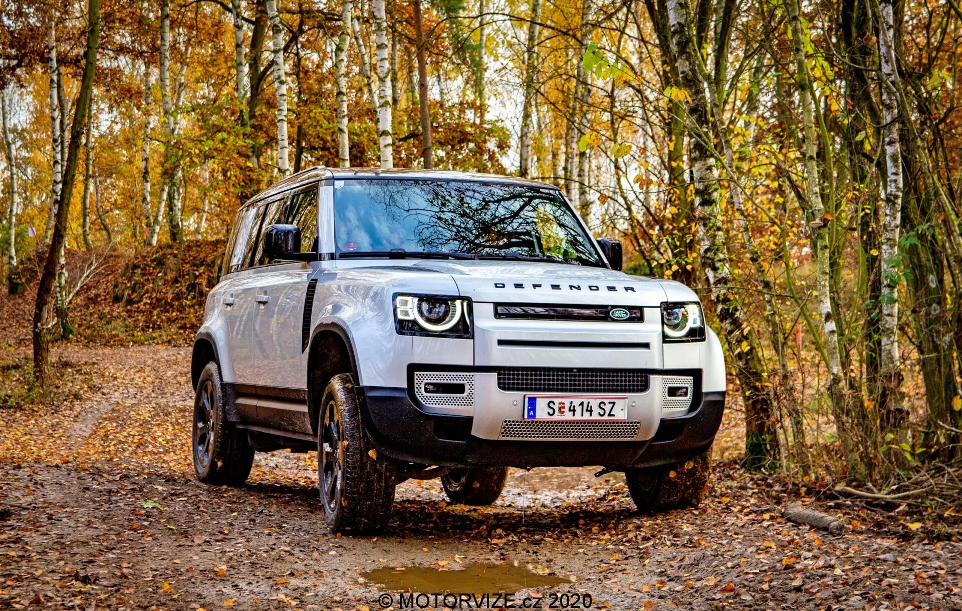 Esta imagem apresenta um SUV Land Rover Defender 2020 posicionado em um caminho florestal não pavimentado, exibindo um design moderno e quadrado a partir de um ângulo de três quartos frontal. A grade, os faróis e o perfil lateral do veículo são claramente visíveis. Os faróis LED distintivos com luzes diurnas, a grade proeminente e os faróis de neblina pronunciados se destacam. Rodas de liga leve grandes com padrão de piso off-road, espelhos laterais com sinais de indicação integrados e uma altura elevada do solo enfatizam sua capacidade off-road. O exterior robusto, as maçanetas tradicionais e a cor neutra da carroceria aumentam ainda mais sua atração como um SUV moderno pronto para aventuras.