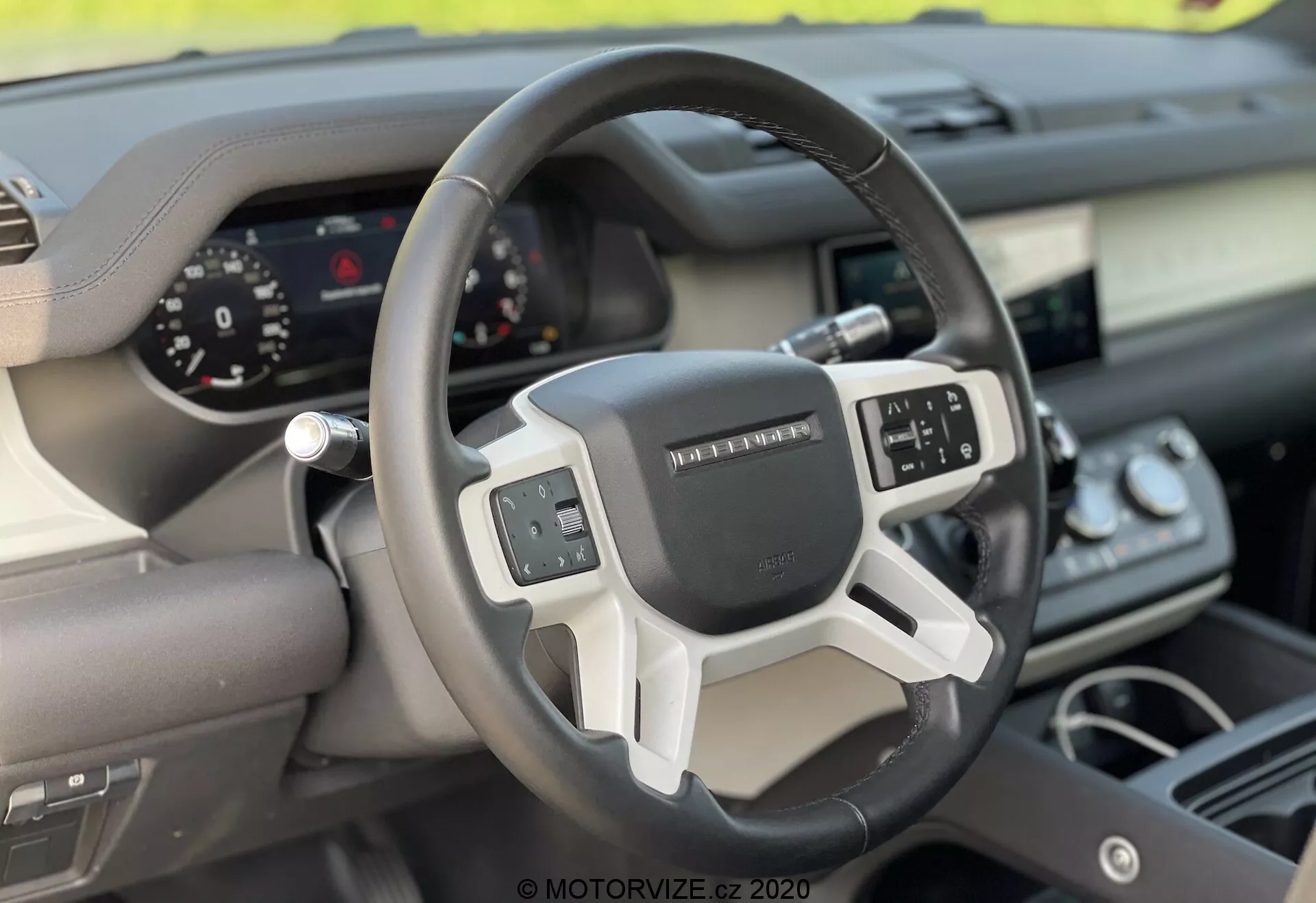 Vista interior do Land Rover Defender 2020 focada no volante e no painel de controlo a partir da perspectiva do assento do passageiro dianteiro. O volante moderno de três raios possui controles multifunções e um painel de instrumentos digital para exibição clara dos indicadores. A combinação de materiais como couro e acabamentos metálicos ou plásticos realça o design ergonômico e tecnológico do veículo.