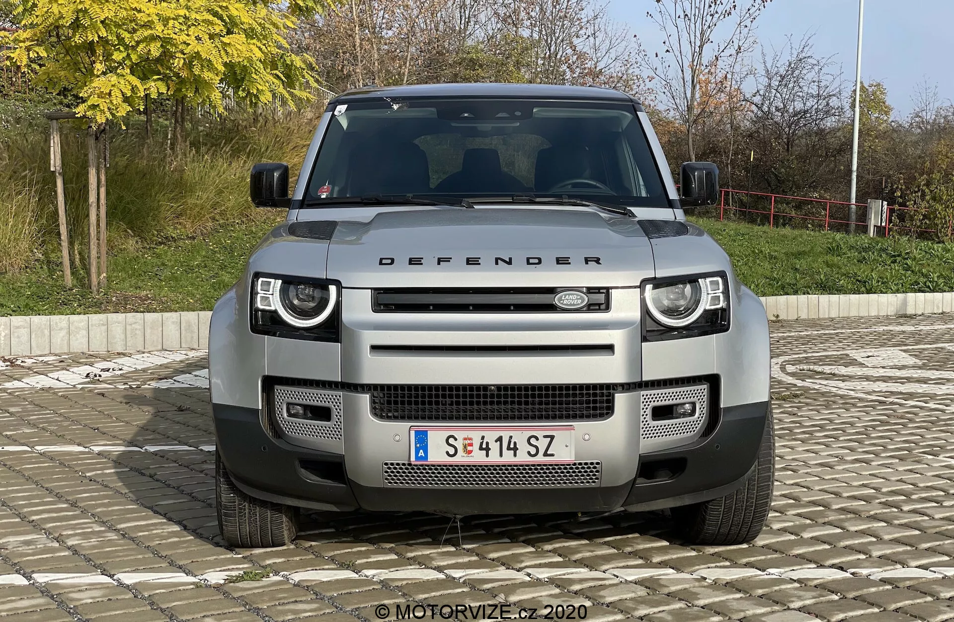Vista frontal do SUV Land Rover Defender 2020, destacando sua grade distintiva, faróis e capô com a inscrição 'DEFENDER'. A imagem centralizada é capturada de um ângulo ligeiramente elevado, destacando os faróis modernos de LED, o padrão geométrico da grade, os sensores da câmera frontal, sensores de estacionamento, placa de proteção dianteira e pontos de reboque. O veículo está posicionado contra um fundo externo simples com arbustos e uma grade, sob luz natural do dia.