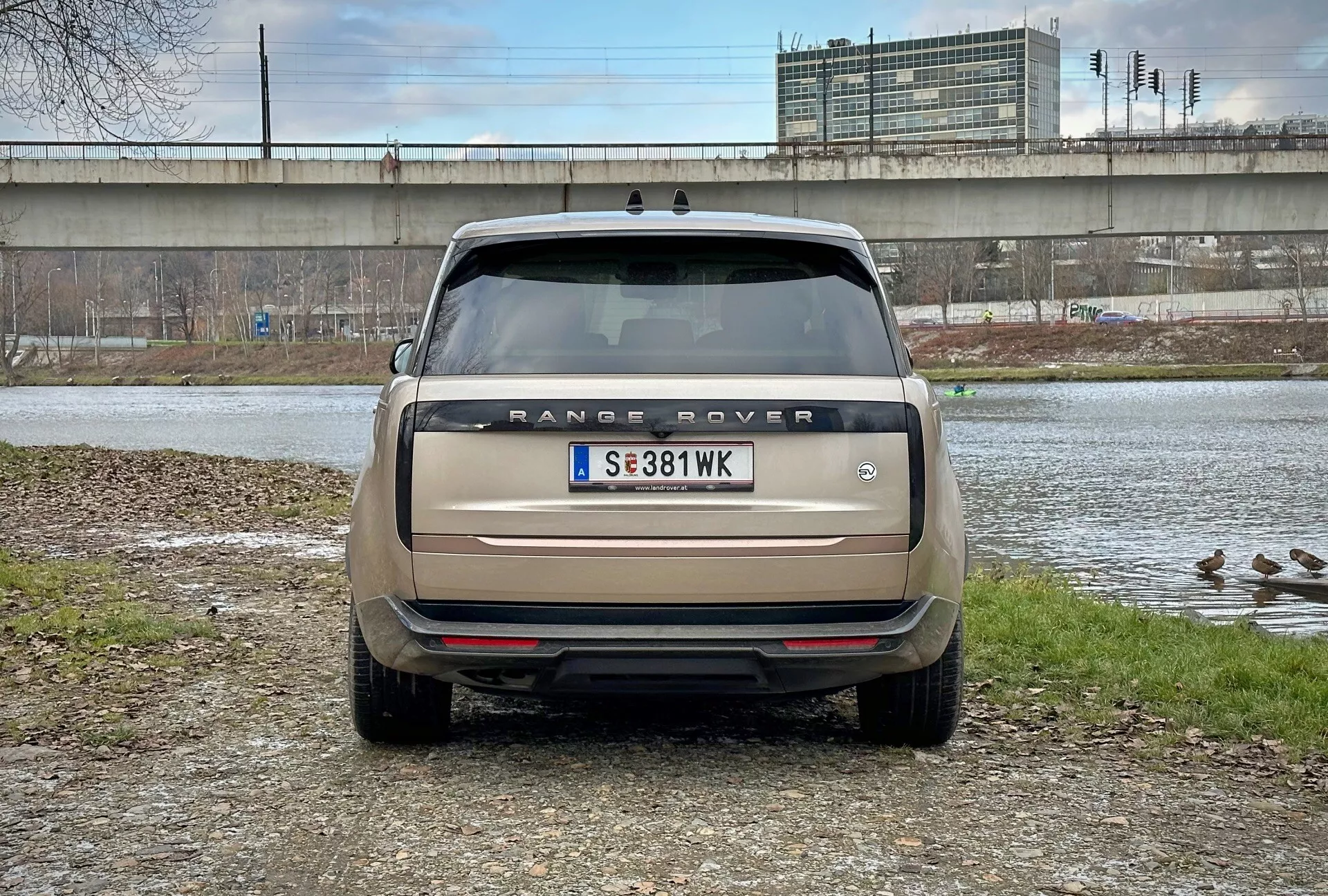 Vista traseira do Range Rover 2021 com ângulo de visão direta ao nível dos olhos, estacionado perto de um corpo de água com patos e um viaduto ao fundo, exibindo o nome do modelo, luzes traseiras, sensores de estacionamento e sistema de escape duplo em bege mate
