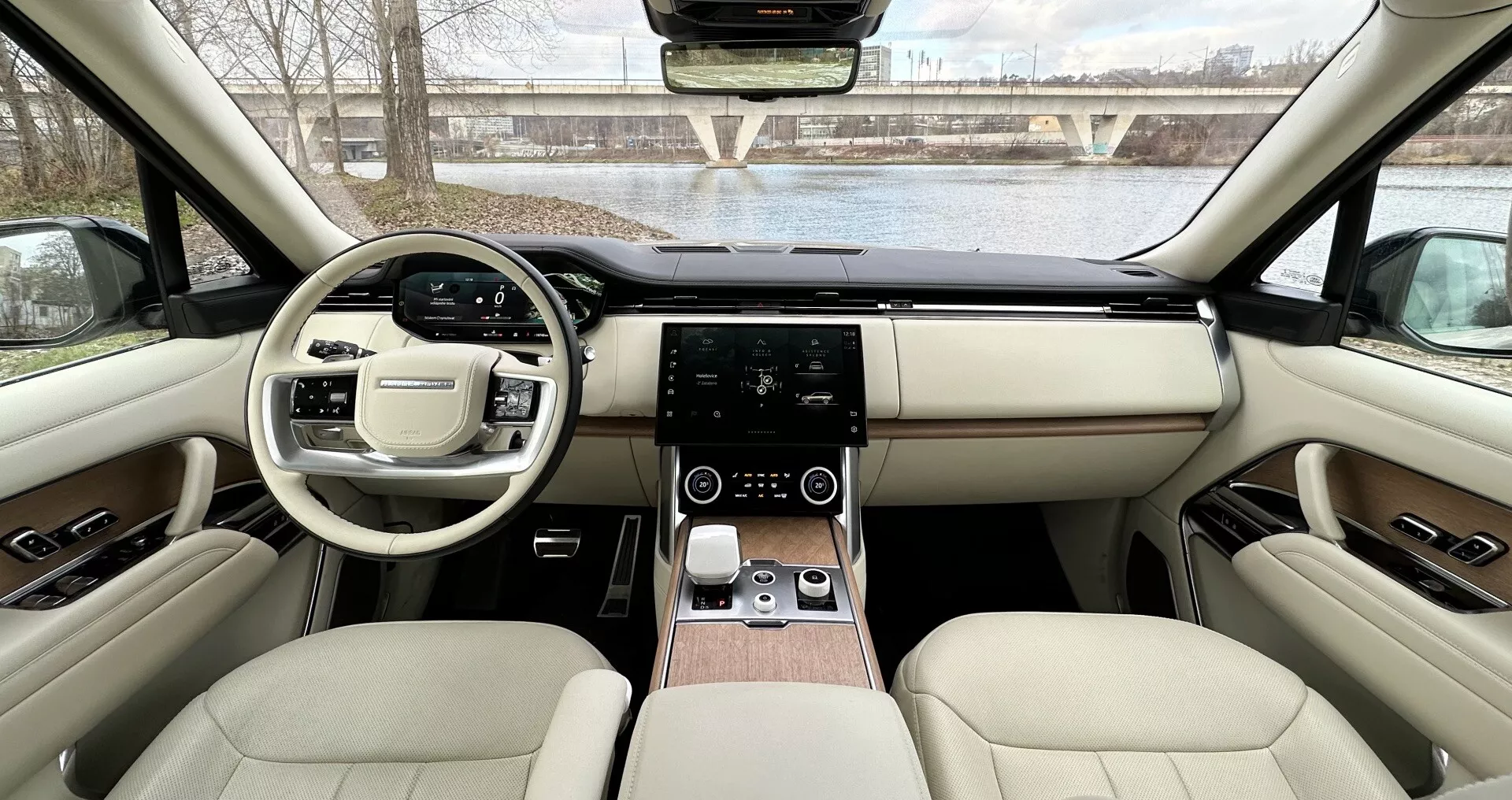 Vista interior do Land Rover Range Rover 2021 a partir dos assentos traseiros em direção ao painel de instrumentos mostrando design simétrico, volante com botões multifuncionais, sistema de infotainment touchscreen, controle de clima digital, estofamento de couro de luxo com detalhes em madeira e características tecnológicas avançadas