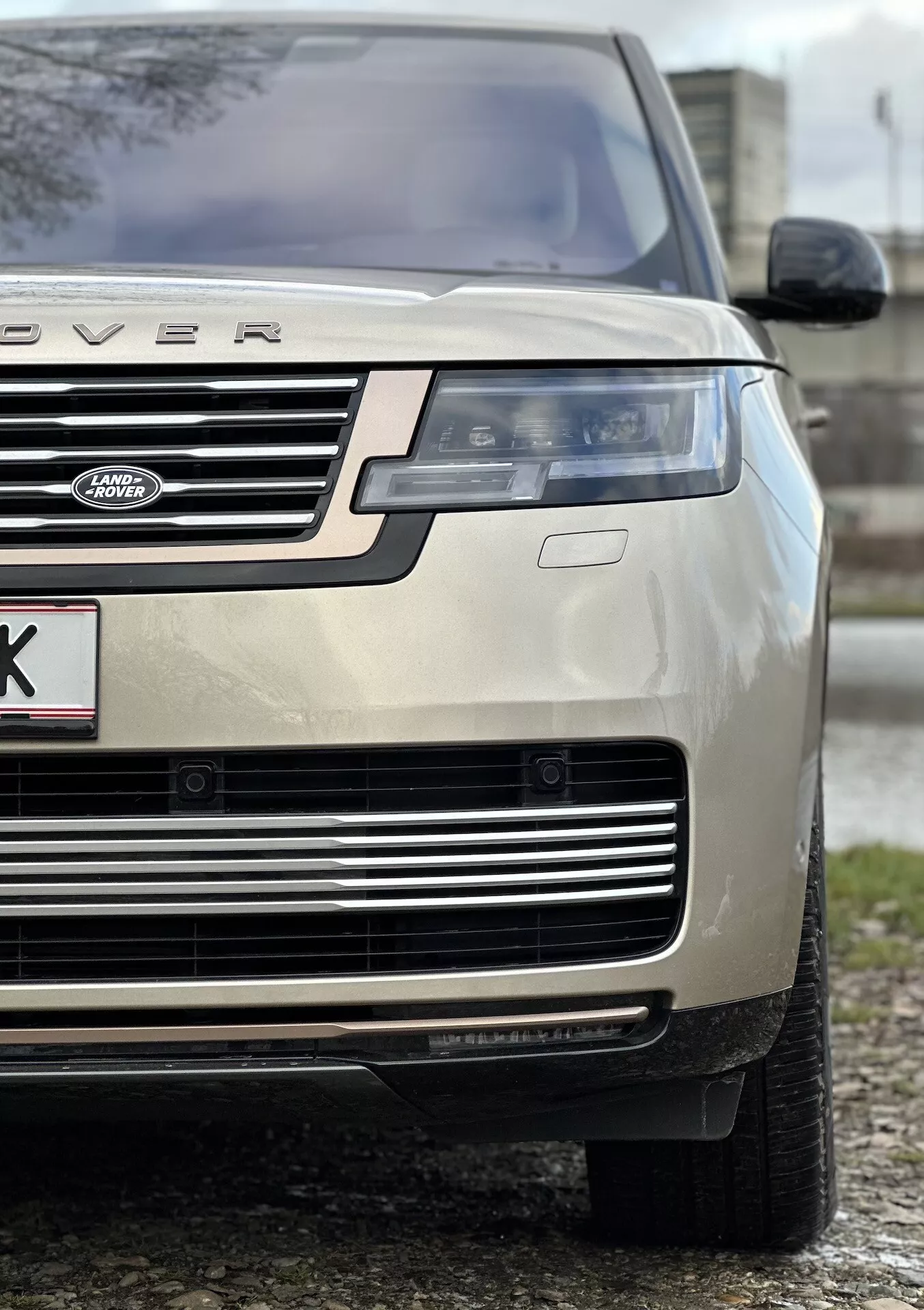 Plano detalhado de um Land Rover Range Rover cor de ouro de 2021 destacando o quarto frontal esquerdo, enfatizando a altura e postura do veículo com foco no capô, grade, farol LED, parachoque e pneu para todas as estações, indicativo de luxo e capacidade off-road.