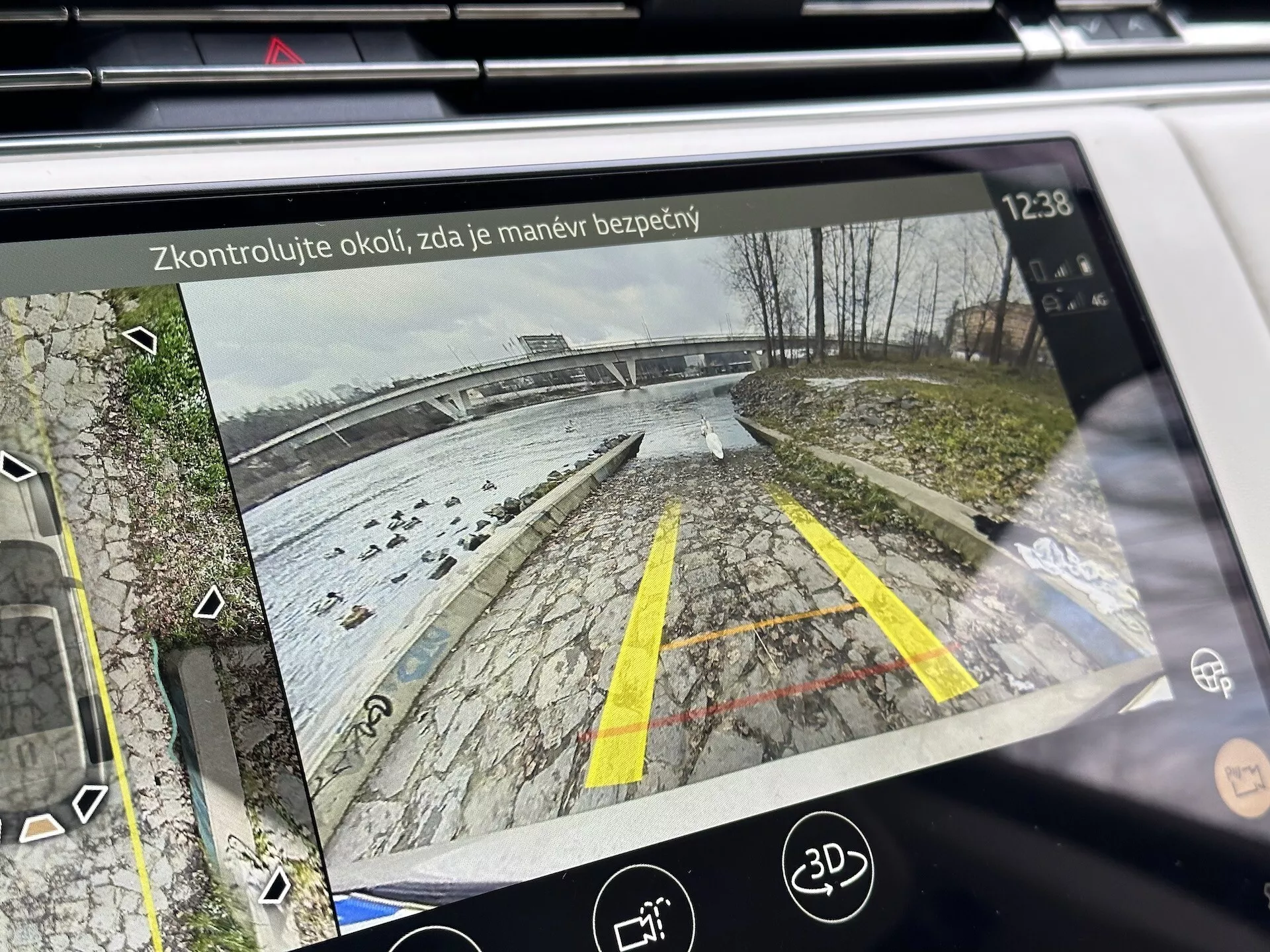 Tela de infotenimento do Land Rover Range Rover 2021 mostrando a vista traseira da câmera de ré com linhas de orientação dinâmicas em uma área de estacionamento perto de uma orla, detalhada com vegetação, ícones de assistência de estacionamento e texto de lembrete de segurança