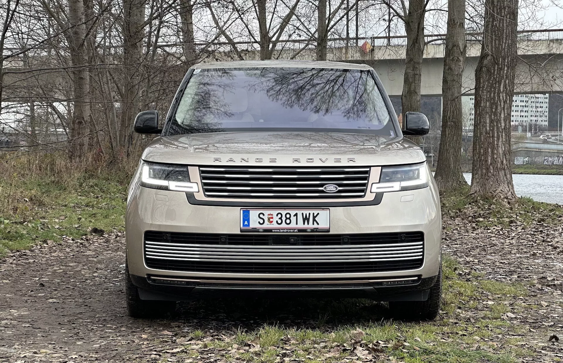 Vista frontal de um Range Rover 2021 mostrando a grelha, faróis LED, a inscrição 'RANGE ROVER' no capô e estacionado numa superfície não pavimentada com um cenário semi-rural