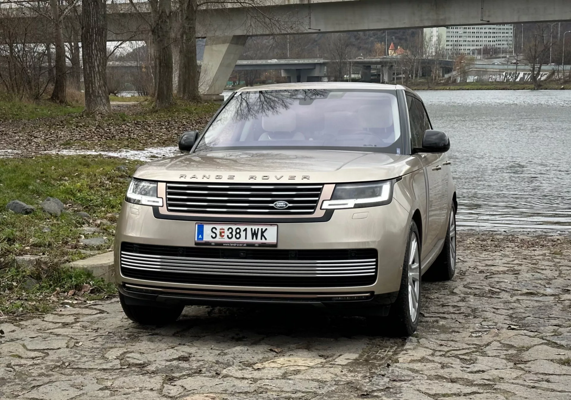 SUV de luxo Range Rover 2021 em prata, destacando a grade frontal proeminente com a marca, luzes diurnas de LED elegantes, para-choque dianteiro com sensores de estacionamento e design de capô clamshell em superfície de paralelepípedos perto de um rio