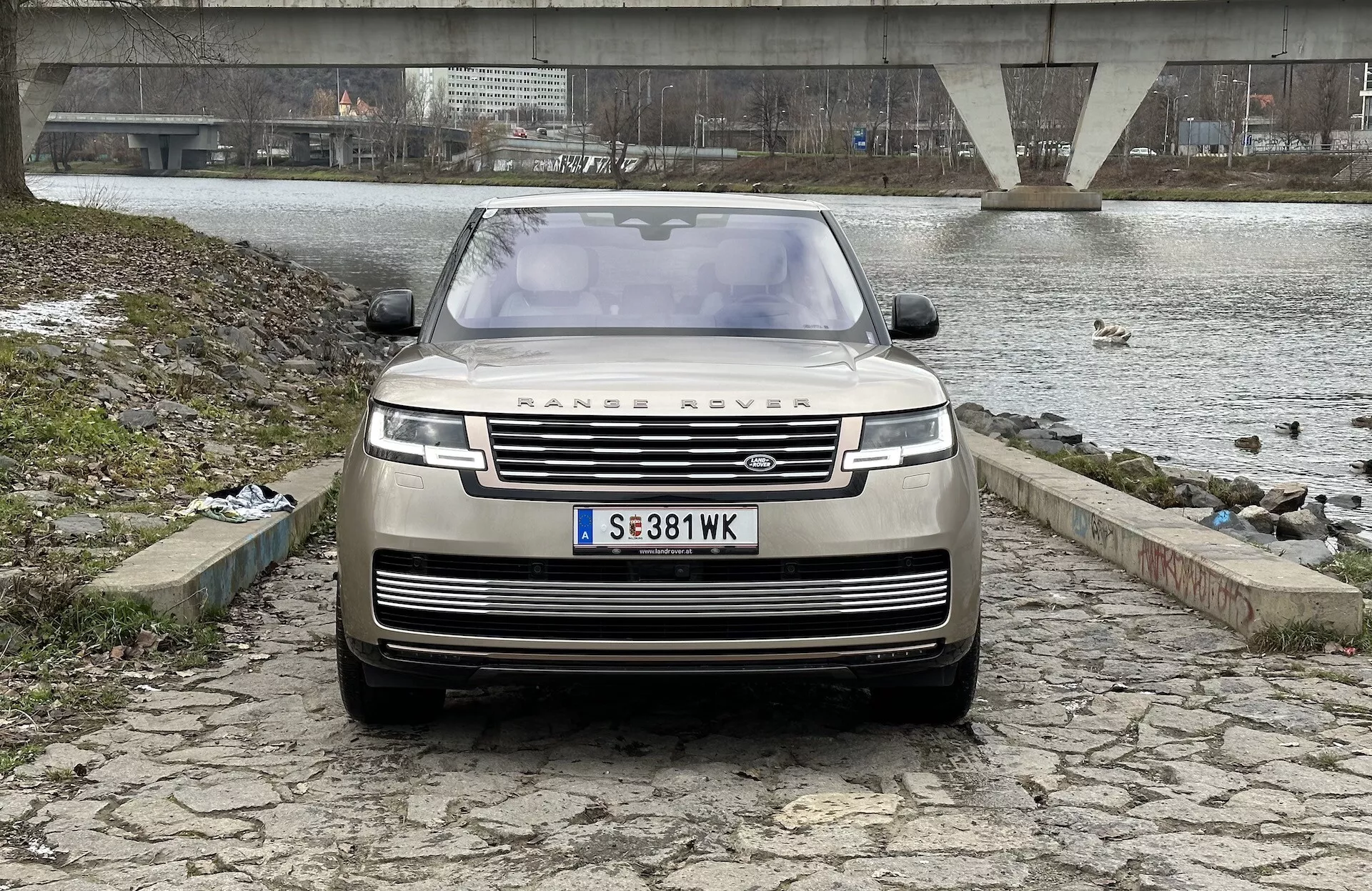 Land Rover Range Rover 2021 fotografado de um ângulo frontal e levemente baixo, enfatizando a grade, para-choque dianteiro e capô, com o emblema icônico RANGE ROVER, faróis LED modernos e tecnologia avançada de assistência ao condutor, contra um fundo de um corpo de água com uma ponte e um céu nublado