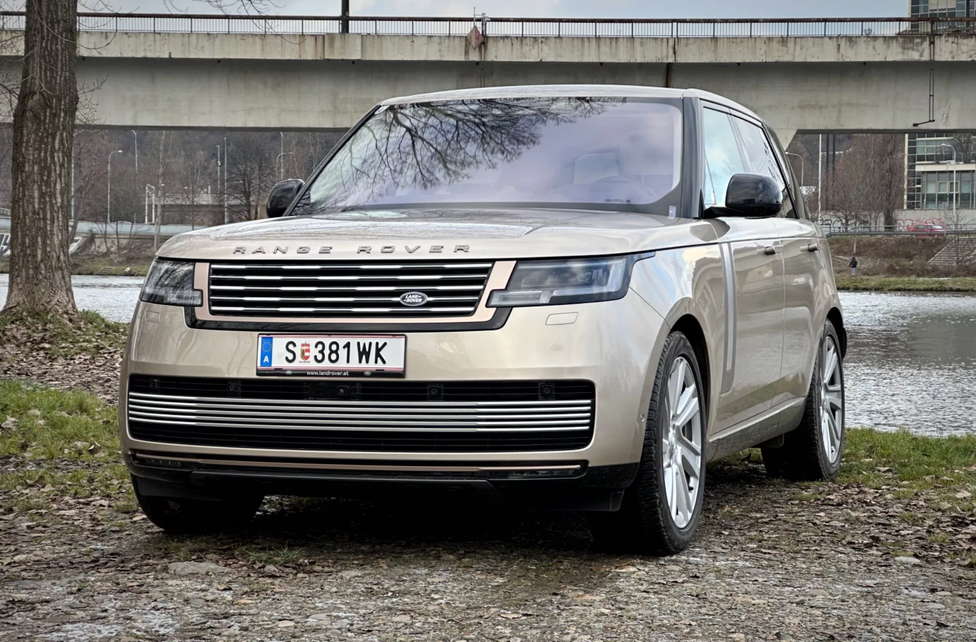 Range Rover de 2021 em uma vista frontal impressionante de três quartos mostrando sua grade distintiva, faróis LED elegantes e placa de proteção frontal robusta contra um cenário de parque urbano, enfatizando luxo e capacidade off-road.