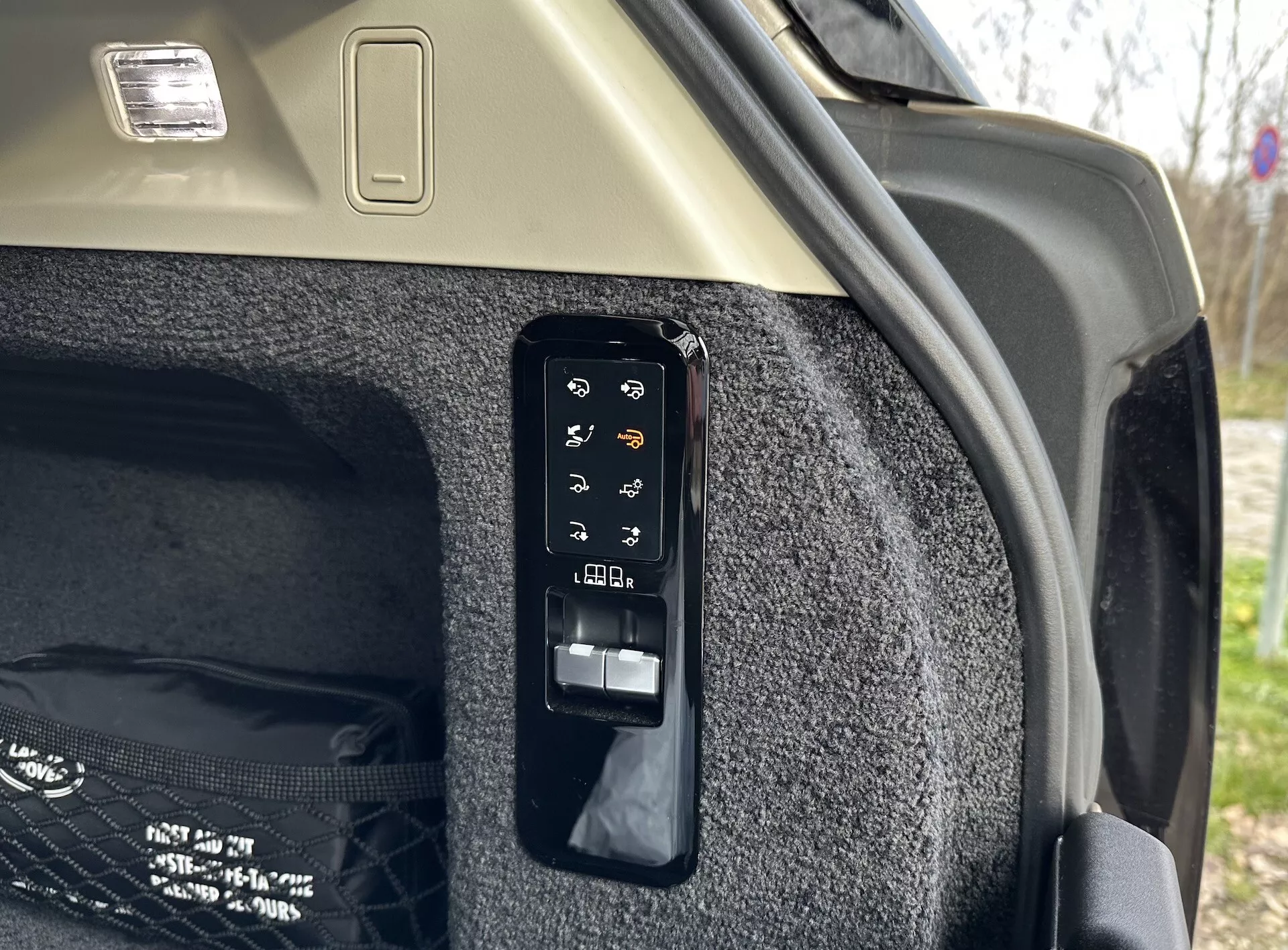 Vista detalhada do painel de controle eletrônico do porta-malas no interior de um Land Rover Range Rover 2021, mostrando botões para operações como abertura e fechamento eletrônicos, controle de cobertura de carga retrátil, possivelmente uma luz de porta-malas ou tomada de energia auxiliar, ajustes dos assentos traseiros a partir do porta-malas e liberação de emergência, situado contra a lateral do porta-malas carpetada e o teto de cor clara com luminária de carga.