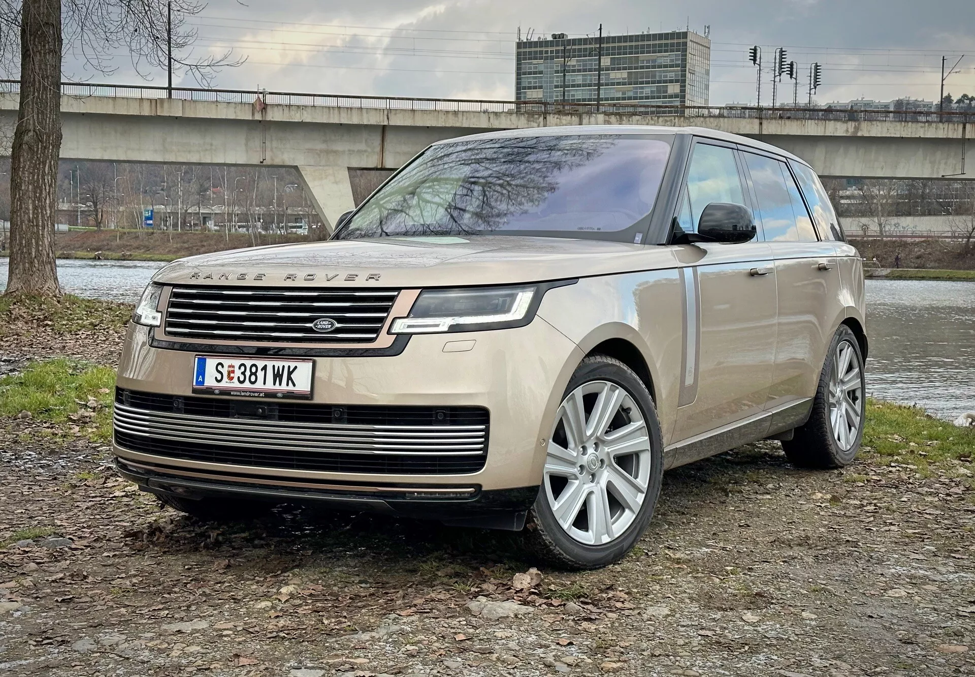 SUV de luxo Land Rover Range Rover 2021 estacionado em terreno misto de terra e grama com uma estrutura de concreto e vegetação ao fundo, mostrando vista do ângulo frontal destacando faróis de LED, rodas de liga e características aerodinâmicas modernas