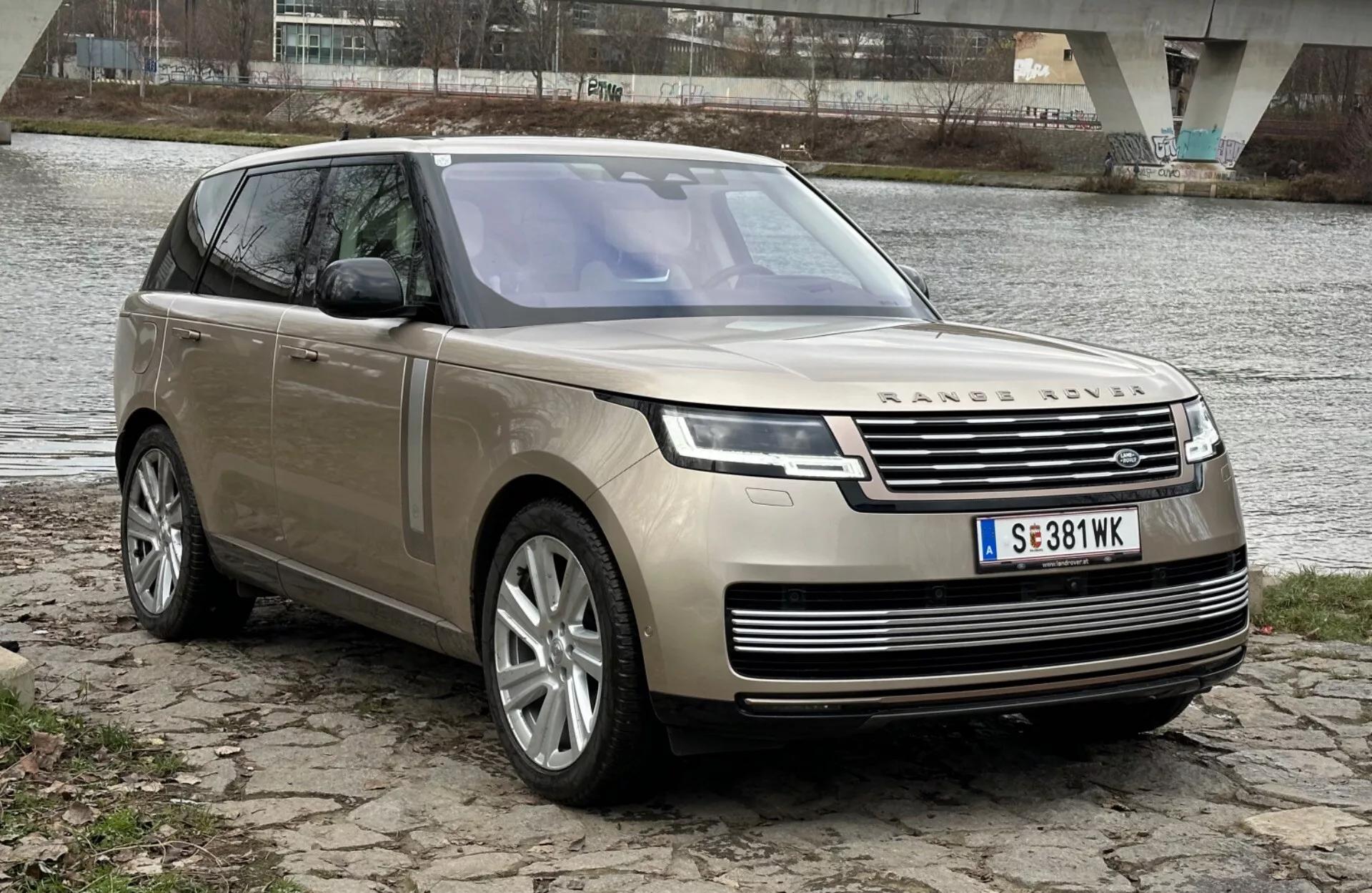 SUV de luxo Land Rover Range Rover 2021 estacionado à beira de um rio com uma ponte ao fundo, exibindo seu design moderno e recursos tecnológicos incluindo faróis de LED, grade frontal com emblema, rodas de liga leve multirraios e teto solar panorâmico.