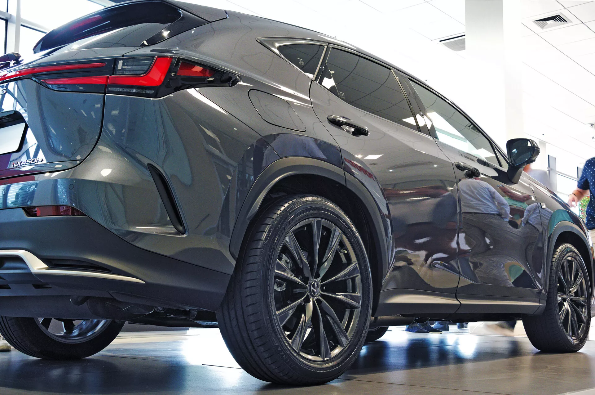 Ângulo traseiro de três quartos do SUV Lexus NX 2021 mostrando o perfil lateral e posterior com grandes rodas de liga leve multirraios, pneus de baixo perfil, luzes traseiras de LED angulares e nítidas, carroceria metálica, janelas escurecidas, spoiler traseiro integrado e ambiente de showroom polido que enfatiza a estética moderna e os recursos de luxo