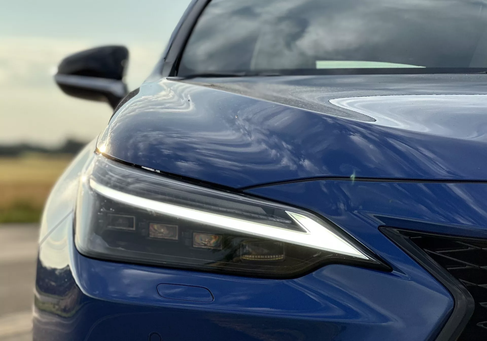 Close-up do quarto dianteiro esquerdo do Lexus NX 2021 caracterizando conjunto de farol LED, capô esculpido, para-choque dianteiro com grade e acabamento azul profundo brilhante, enfatizando a tecnologia de iluminação avançada e design aerodinâmico.