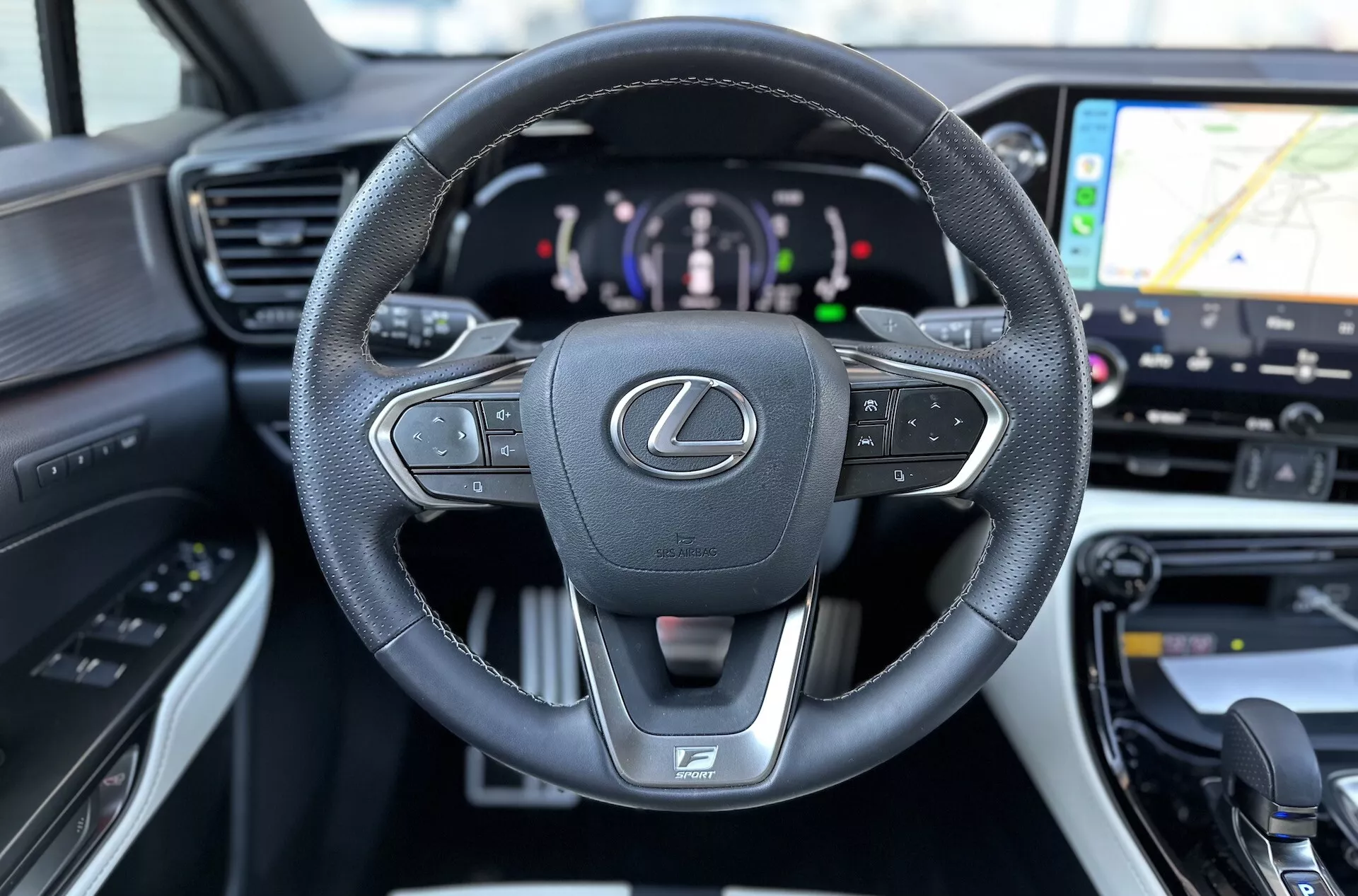 Interior do Lexus NX 2021 focando no volante multifuncional revestido de couro com emblema Lexus e logótipo F SPORT, painel de instrumentos digital indicando sistema de propulsão híbrida e vislumbre do sistema de navegação GPS no sistema de infotenimento
