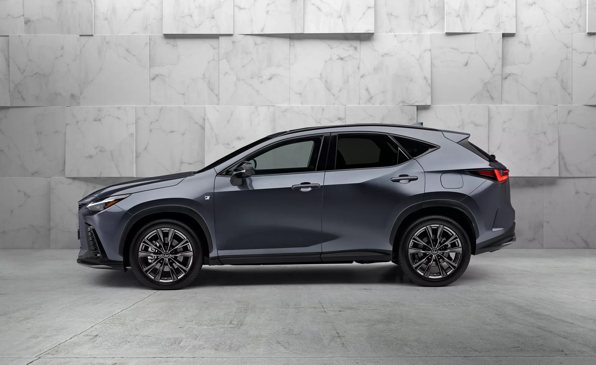Vista lateral de um moderno SUV crossover Lexus NX 2021, exibindo sua maior distância ao solo e forma da carroceria. Pintado em uma tonalidade escura, possivelmente cinza ou azul, o veículo possui cinco portas, design aerodinâmico elegante, grandes rodas de liga leve multiraio, faróis e lanternas traseiras finos em LED, espelhos laterais na cor da carroceria com indicadores de seta integrados, janelas com acabamento cromado e uma antena em forma de barbatana de tubarão, tudo isso em um fundo de mármore.