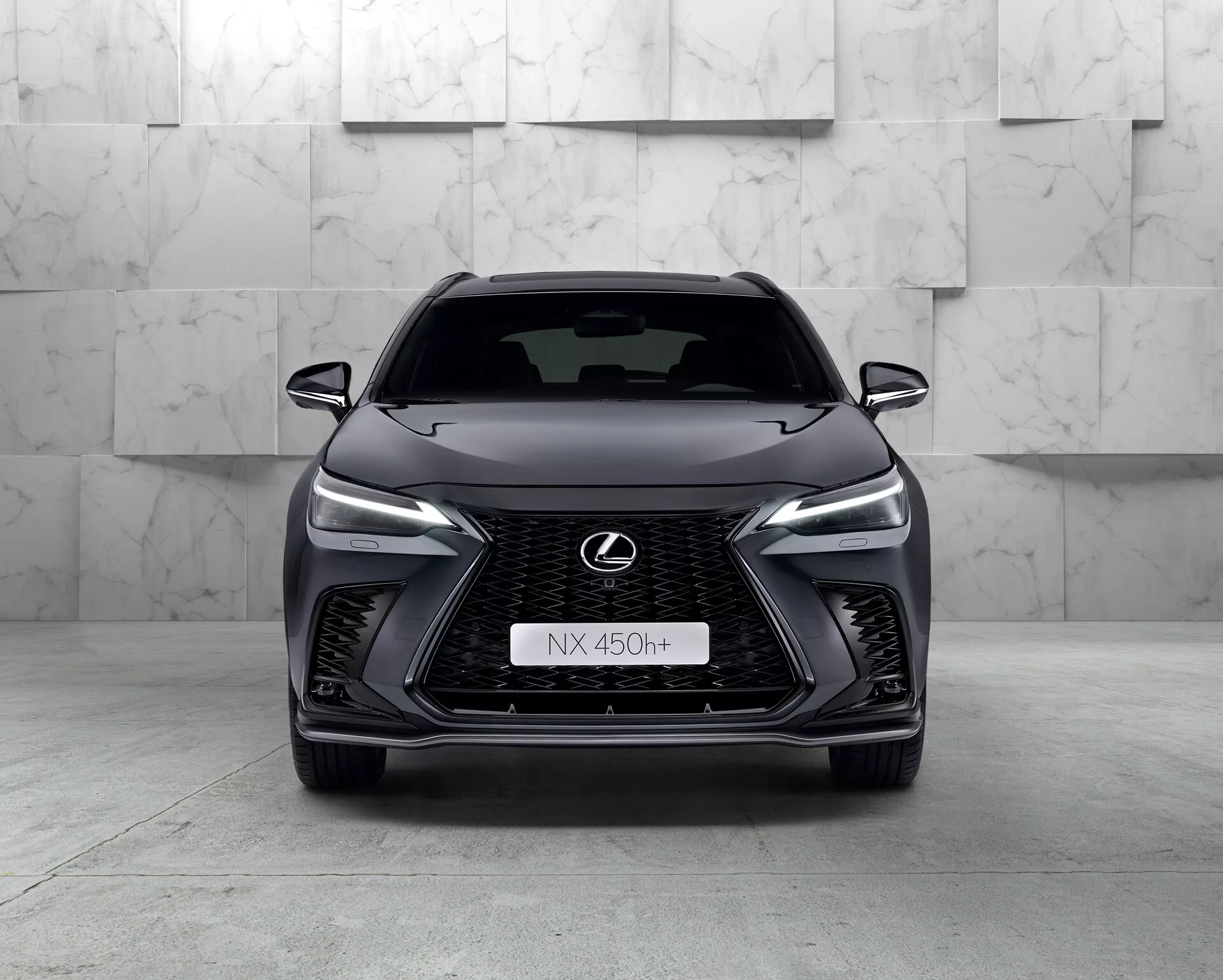 Vista frontal do Lexus NX 450h+ 2021, um SUV moderno e elegante que exibe sua proeminente grade em forma de fuso com padrão de malha, faróis de LED estreitos, emblema da Lexus cromado com contorno azul e design agressivo do para-choque dianteiro. A natureza híbrida do carro é indicada pelo emblema 'h+' na grade. O ambiente do estúdio destaca as curvas e características do veículo com iluminação limpa e controlada.