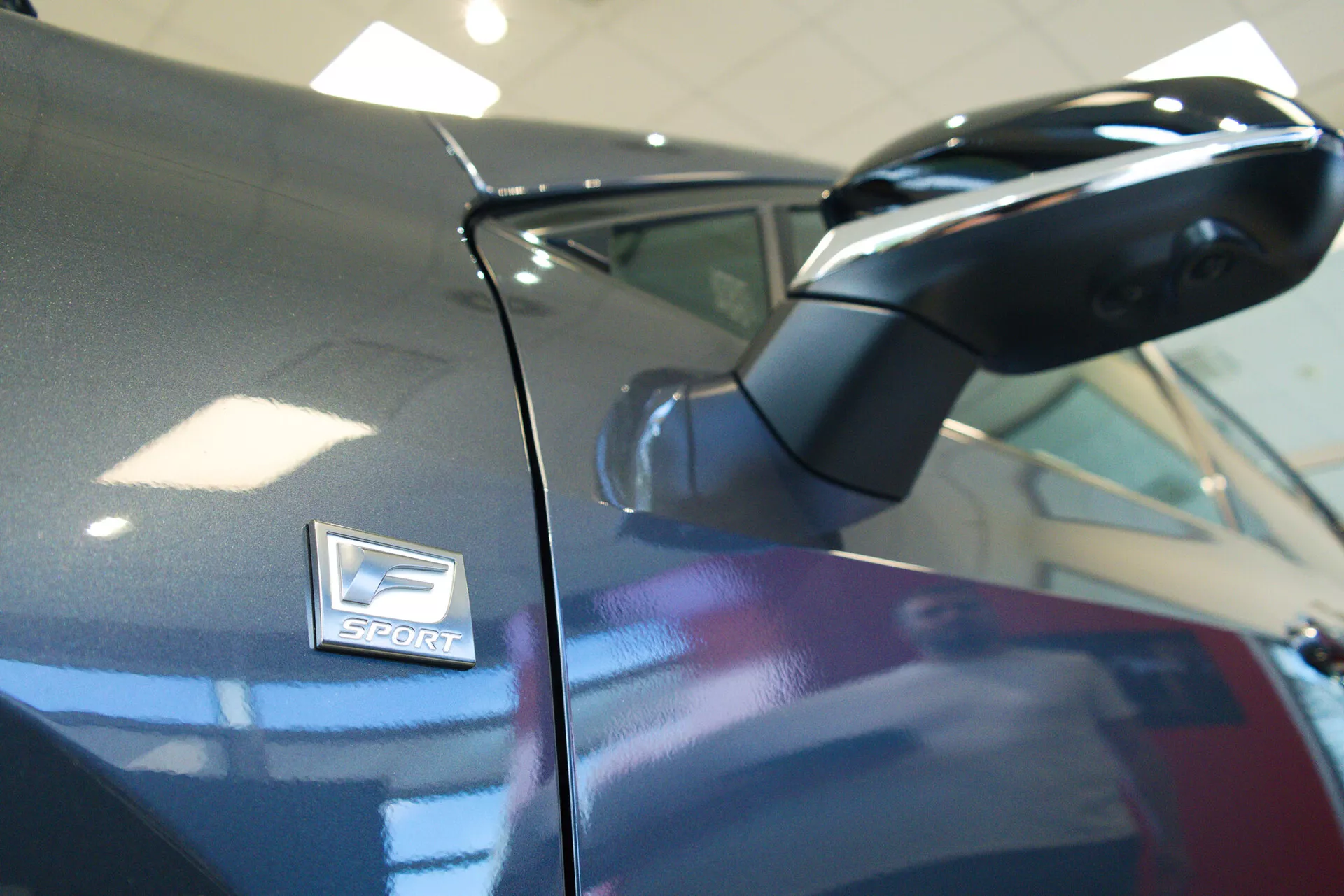 Fotografia em close-up de um Lexus NX 2021, focando no espelho lateral e no emblema 'Sport'. A foto, tirada de um ângulo baixo levemente inclinado para cima, destaca o espelho aerodinâmico na carroceria de cor escura. O emblema 'Sport', indicativo de uma variante orientada para desempenho, está posicionado acima do arco da roda com uma profundidade de campo rasa que realça a estética do design.