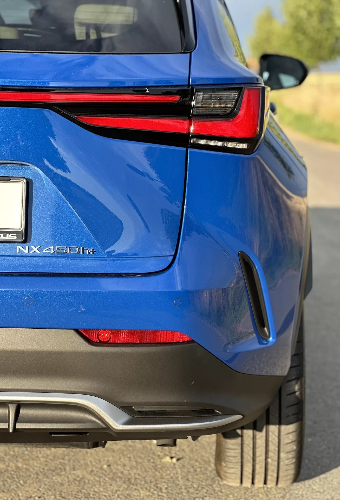 Visão angular aproximada da seção traseira de um SUV crossover híbrido Lexus NX 450h+ azul, destacando seu acabamento brilhante, luzes traseiras LED, distintivo híbrido e características de design esportivo