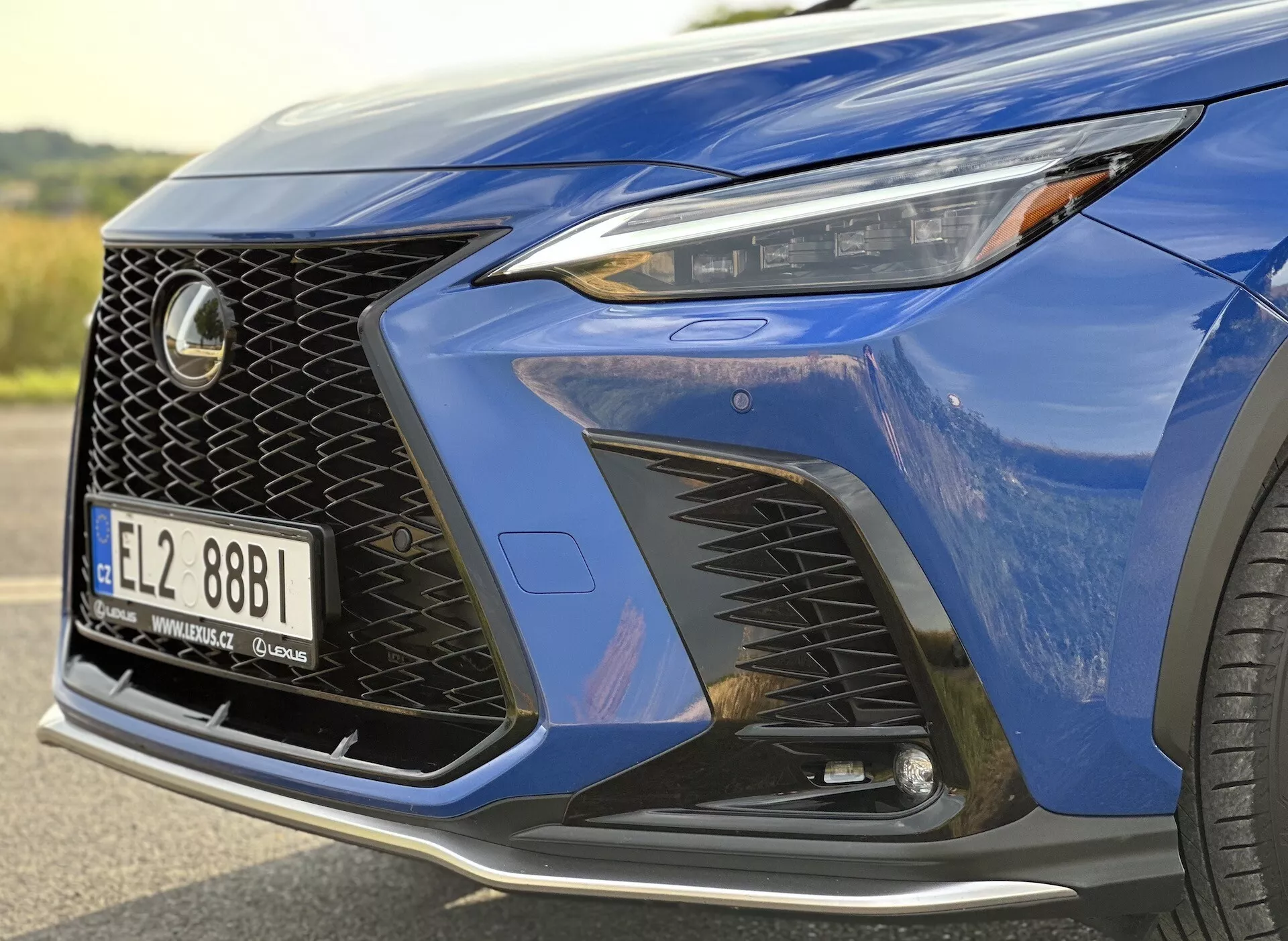 Close-up do canto frontal do Lexus NX 2021 destacando o farol com tecnologia LED, design distintivo da grade em malha e para-choque dianteiro aerodinâmico com entradas de ar, realçado por uma pintura metálica azul profundo e um vislumbre de uma roda de liga luxuosa.