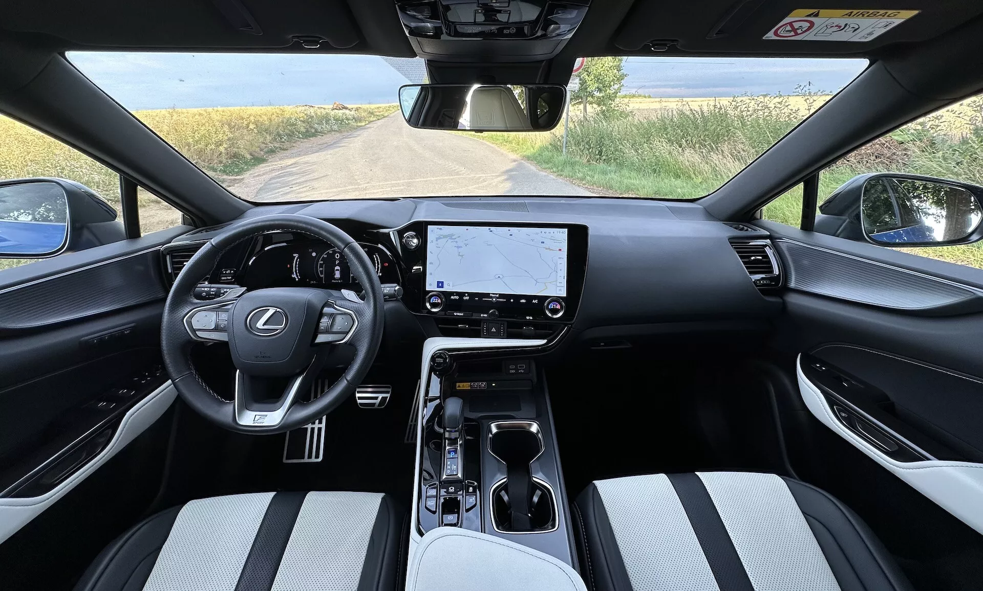 Vista interior do Lexus NX 2021 do ponto de vista do motorista, exibindo um volante centralizado com logo da Lexus e botões de controle, um grande touchscreen de infotainment no painel, controles climáticos minimalistas, um seletor de mudanças automático no console central com porta-copos e compartimento de armazenamento, assentos estofados em material de duas cores, e uma vista parcial de uma paisagem rural através do para-brisa.