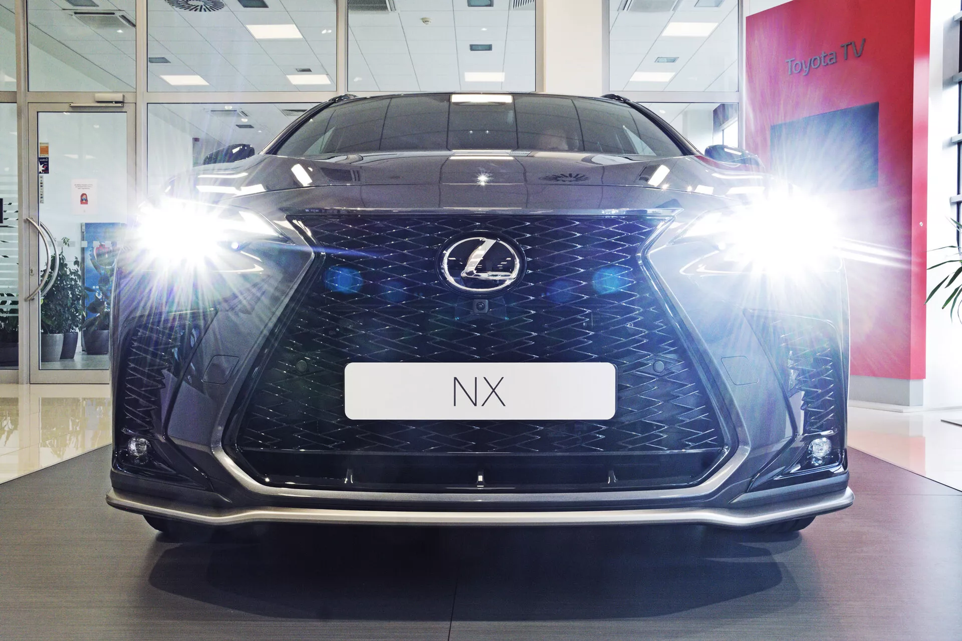 Vista frontal do veículo Lexus NX 2021 fotografado de um ângulo baixo, mostrando sua altura e presença na estrada em um ambiente de showroom. A imagem destaca a grade distintiva em forma de ampulheta com padrão de malha escura, os faróis LED angulares iluminados com efeitos de flare, luzes diurnas e detalhes do para-choque inferior. O chão polido e o suporte de placa com a inscrição 'NX' adicionam ao apelo luxuoso e tecnológico do carro.
