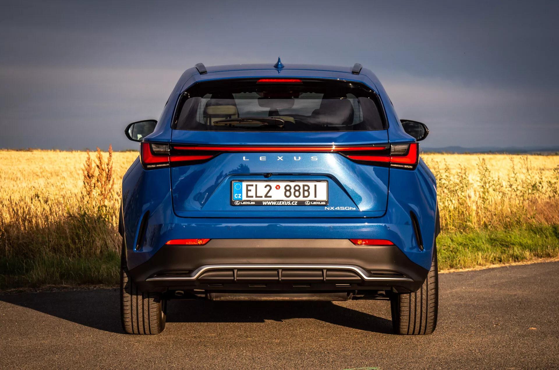 Vista traseira do SUV azul Lexus NX 2021 em estrada de asfalto com campo dourado ao fundo, destacando a tecnologia híbrida e características de design