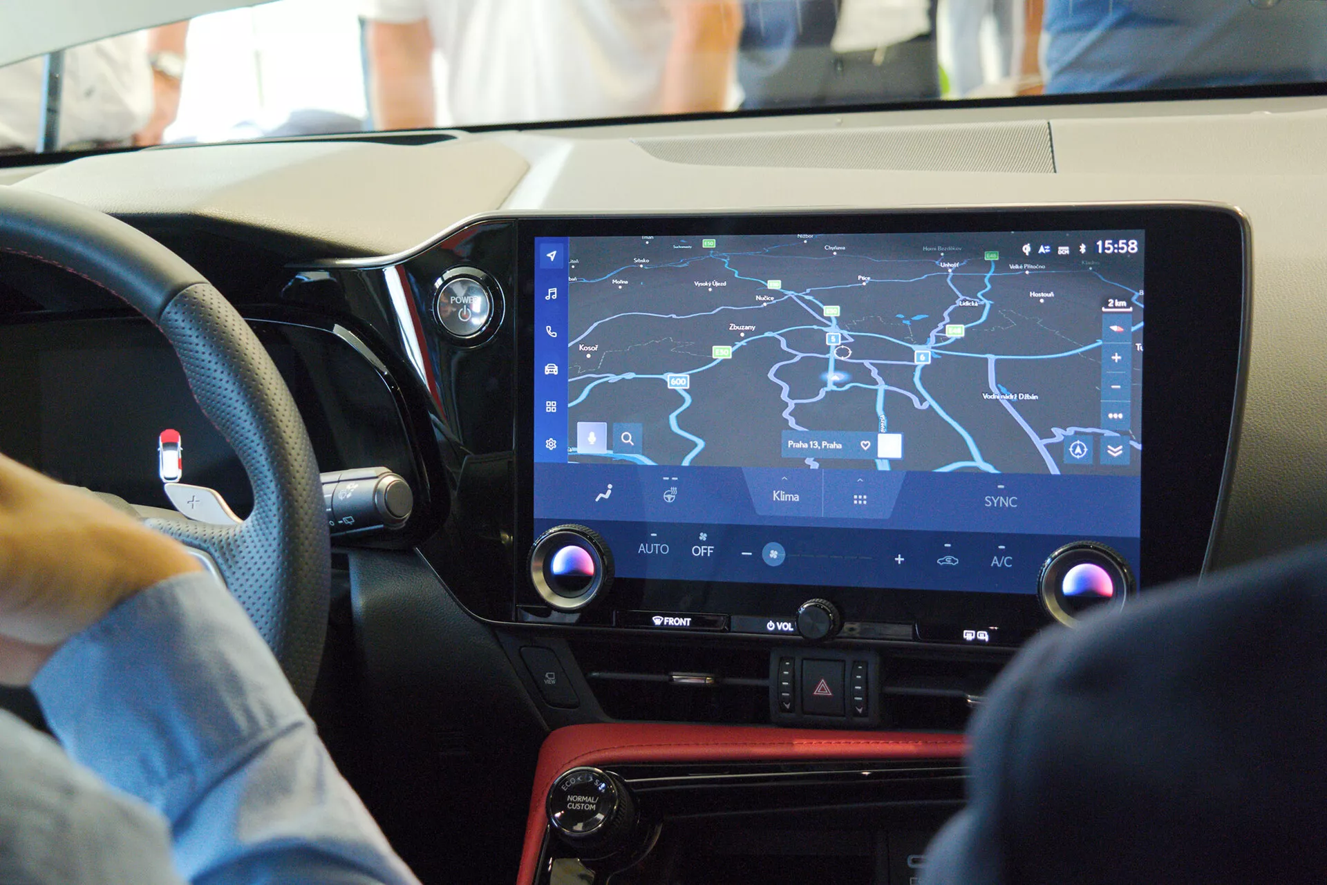 Vista interior de um carro moderno focando na consola central e no sistema de infoentretenimento da perspetiva do passageiro, apresentando um grande ecrã tátil com um mapa de navegação, controlos físicos para funções de climatização, um painel preto brilhante com um detalhe vermelho, e sugerindo um design amigável ao condutor em um ambiente movimentado.