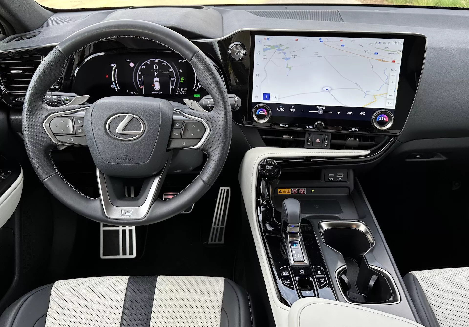 Vista interior do Lexus NX 2021 mostrando a área do motorista e o console central de um ângulo elevado, com o logotipo da Lexus no volante com controles montados, um cluster de instrumentos digitais iluminado, um grande sistema de infotainment touchscreen com navegação GPS, botões de controle de clima e áudio, seletor de marchas ergonômico, controles de modo de condução, materiais luxuosos nos assentos e pontos de contato, acentos metálicos, e porta-copos visíveis e compartimento de armazenamento.