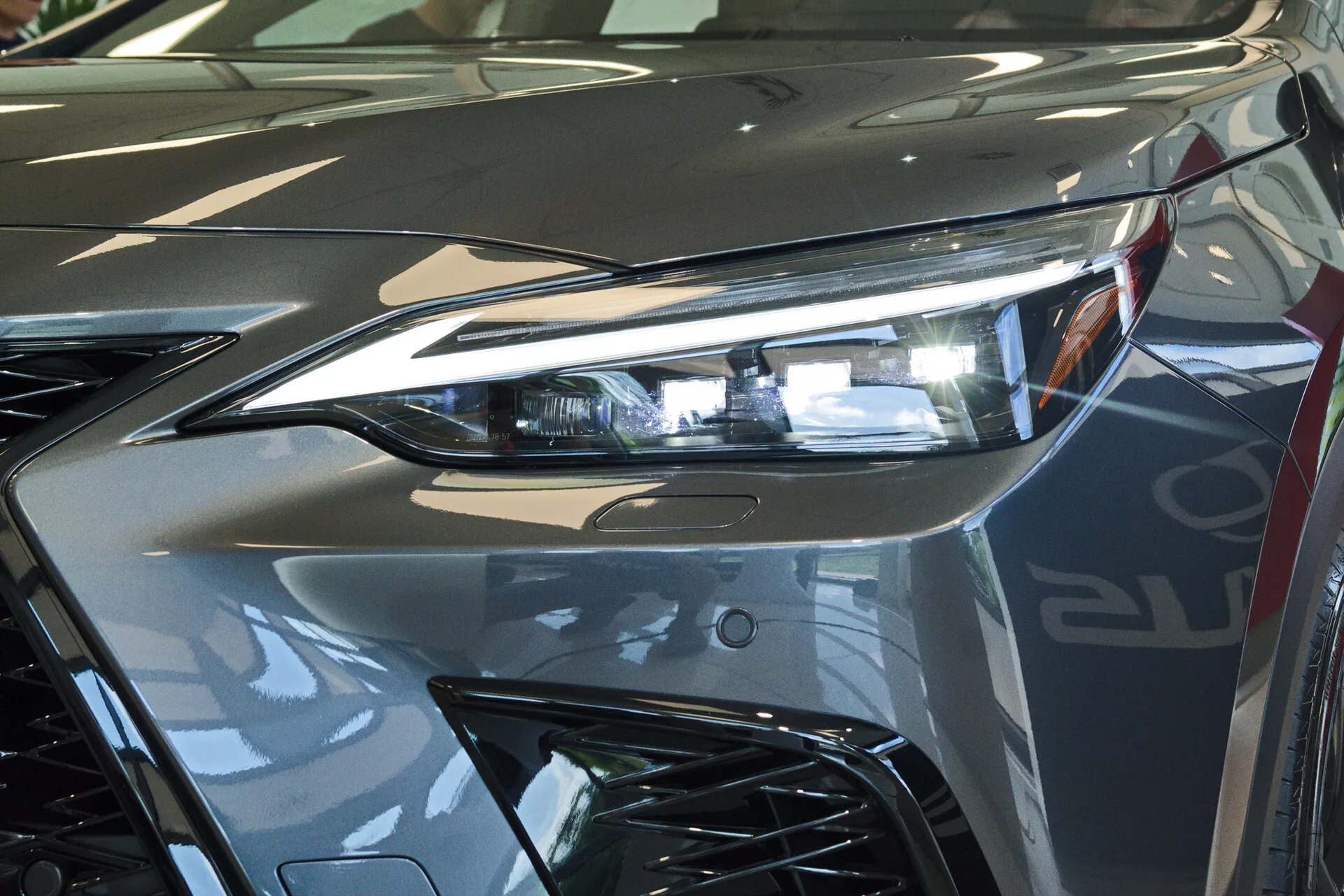 Close-up da parte frontal de um Lexus NX 2021, focando-se na montagem do farol do lado do condutor com tecnologia avançada de iluminação LED e parte da grade de estilo agressivo. A imagem capta o brilho reflexivo da pintura metálica prateada e as linhas dinâmicas que contribuem para a estética aerodinâmica do veículo. Fotografado em um ambiente interno, provavelmente em um showroom, enfatizando o design luxuoso e o acabamento de alta qualidade do veículo.