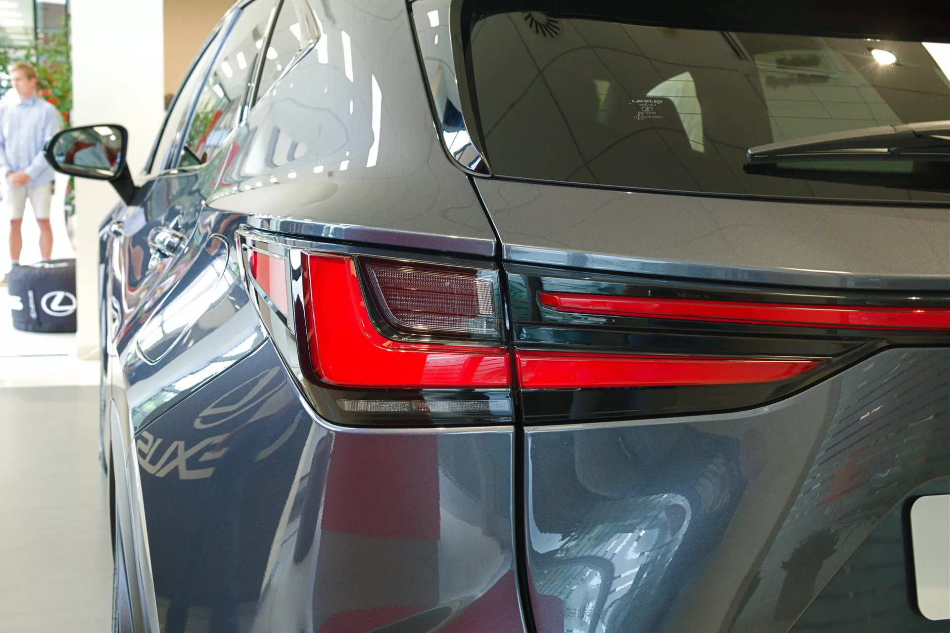 Close-up do Lexus NX 2021 de um ângulo traseiro de três quartos, destacando o design elegante da lanterna traseira em LED, parte do painel traseiro e uma porção da tampa do porta-malas. A imagem mostra a pintura escura e brilhante do carro, as linhas aerodinâmicas do corpo e sugere um ambiente interno de showroom com reflexos de outro veículo, elementos arquitetônicos e uma pessoa caminhando ao fundo.