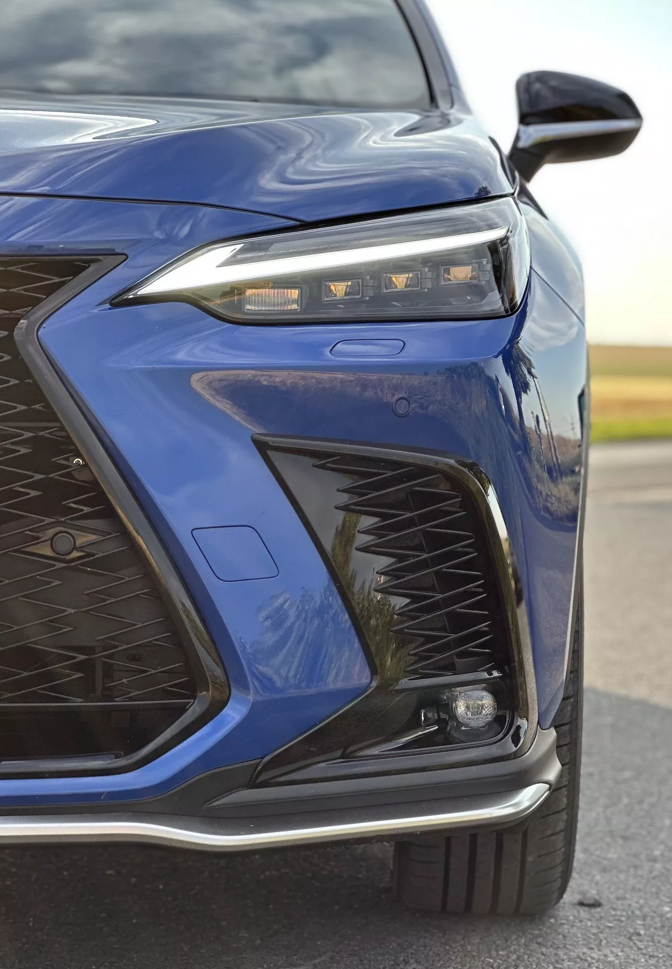 Close-up do canto frontal esquerdo de um Lexus NX 2021 em azul escuro destacando o design avançado de faróis LED, padrão intrincado da grelha, para-choque dianteiro esportivo e possíveis características adaptativas de iluminação e aerodinâmicas com um acabamento metálico brilhante