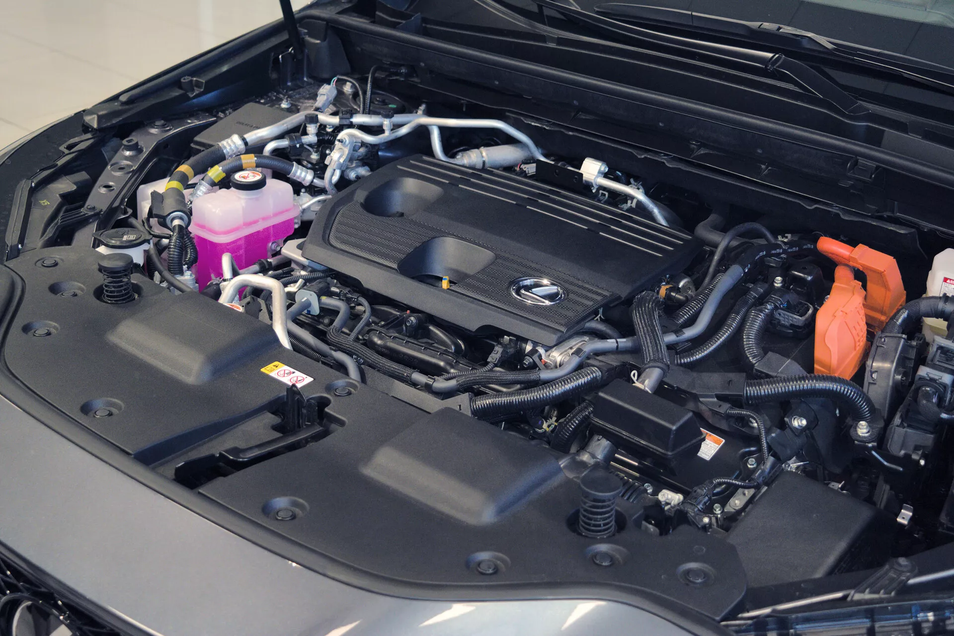 Vista aproximada do compartimento do motor de um carro com o capô aberto, revelando um compartimento de motor bem organizado e limpo. A tampa do motor apresenta um logotipo proeminente, indicando a marca, e vários componentes, como reservatórios coloridos, cabos fixos e uma bateria visível com uma tampa de terminal positivo vermelha. O arranjo sugere um veículo moderno e possivelmente luxuoso, destacando a complexidade do seu sistema elétrico e a condição geral impecável.