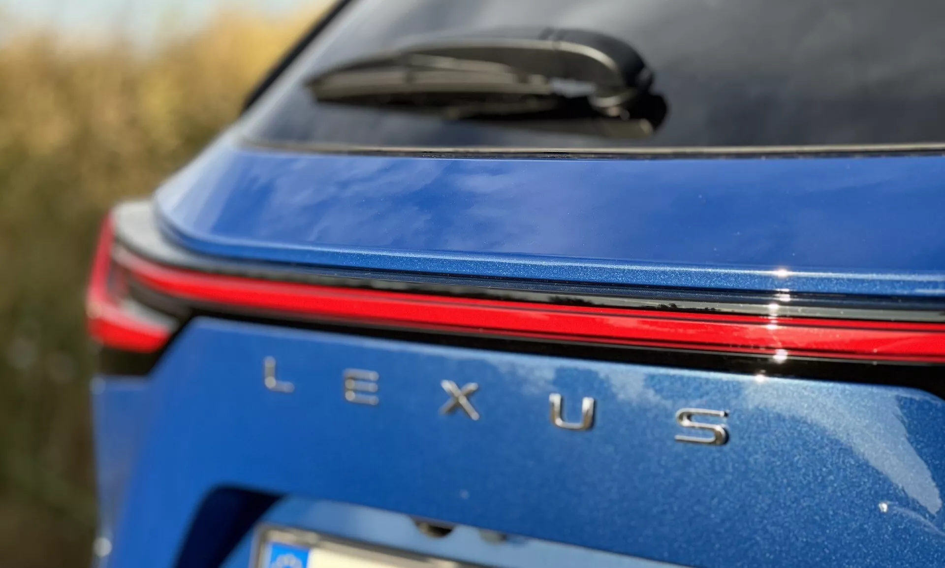 Visão detalhada da área de marcação traseira do Lexus NX 2021 com um distintivo Lexus prateado, uma faixa de luz vermelha acima do nome, em um corpo azul metálico sob condições de luz do dia