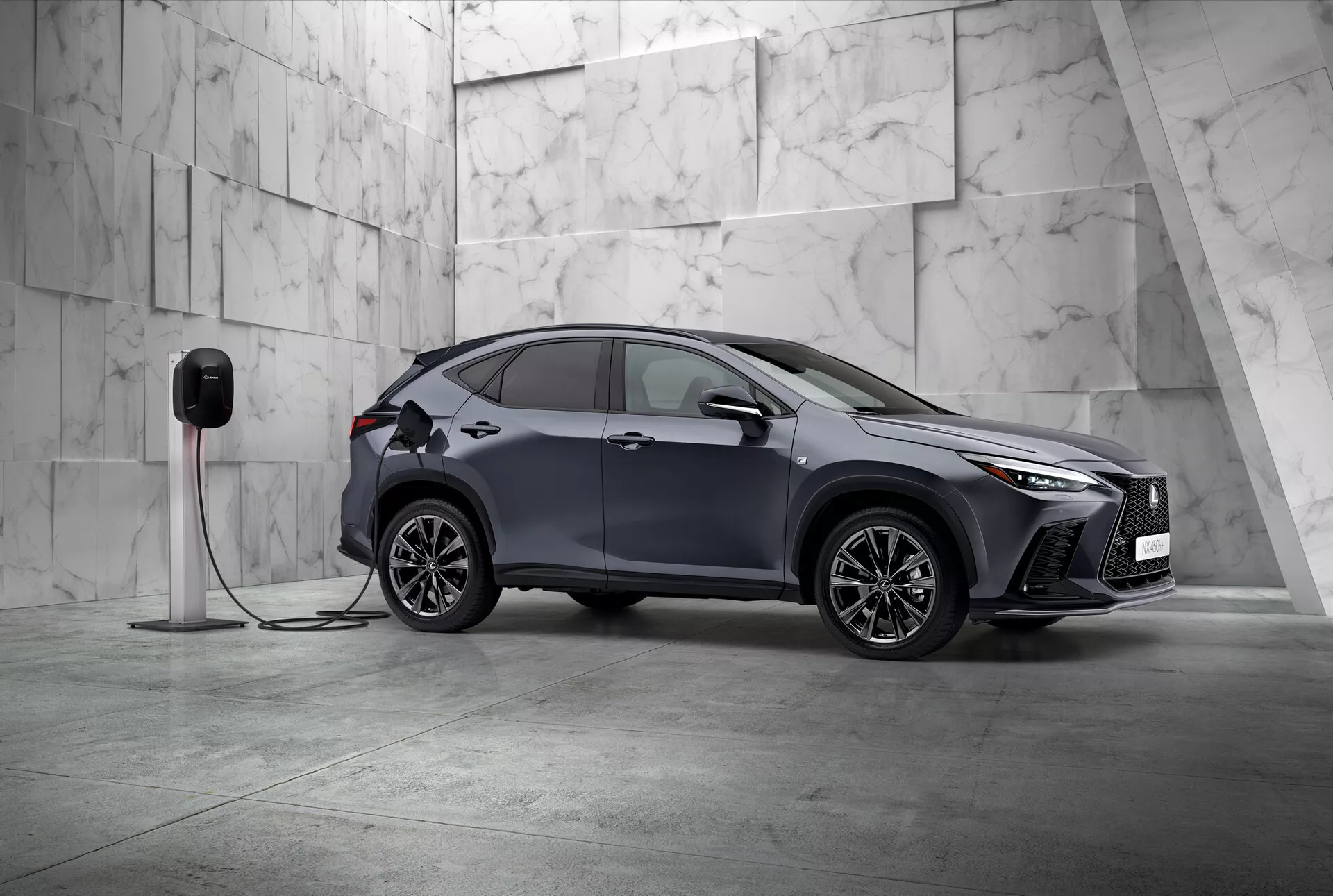Esta imagem apresenta o Lexus NX 2021, um moderno SUV compacto com um design elegante e esportivo, fotografado de uma vista frontal de três quartos. O lado esquerdo do veículo e parcialmente sua frente são visíveis, posicionados diagonalmente em um cenário minimalista adornado com grandes ladrilhos de mármore claro, sugerindo uma atmosfera premium. O Lexus NX exibe faróis LED angulares distintos e uma grade frontal proeminente, enfatizando sua natureza sofisticada. Espelhos laterais aerodinâmicos visíveis, projetados intrincados de liga multi-raios e maçanetas embutidas destacam o design esportivo e avançado. Um detalhe crucial é a conexão do carro a uma estação de carregamento elétrico, sinalizando que é um veículo elétrico ou híbrido plug-in. O para-lama dianteiro inclui um porto de carregamento, indicando suas capacidades elétricas. Esta imagem estática enfoca a exibição do design do veículo e características tecnológicas em uma composição calma e equilibrada com muito espaço negativo, exemplificando a exclusividade e inovações no design automotivo.
