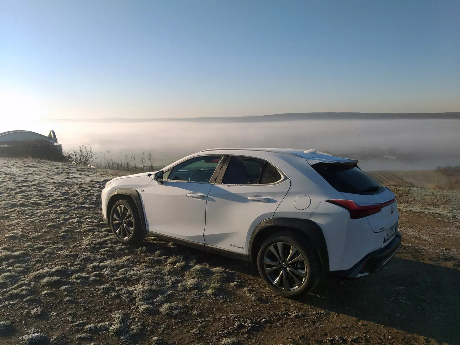 SUV crossover Lexus UX 2019 estacionado em grama coberta de geada com fundo de paisagem nebulosa, exibindo o perfil do veículo de um ângulo traseiro de três quartos destacando linhas de caráter lateral, arcos de roda, linha do teto afinando e rodas de liga leve.