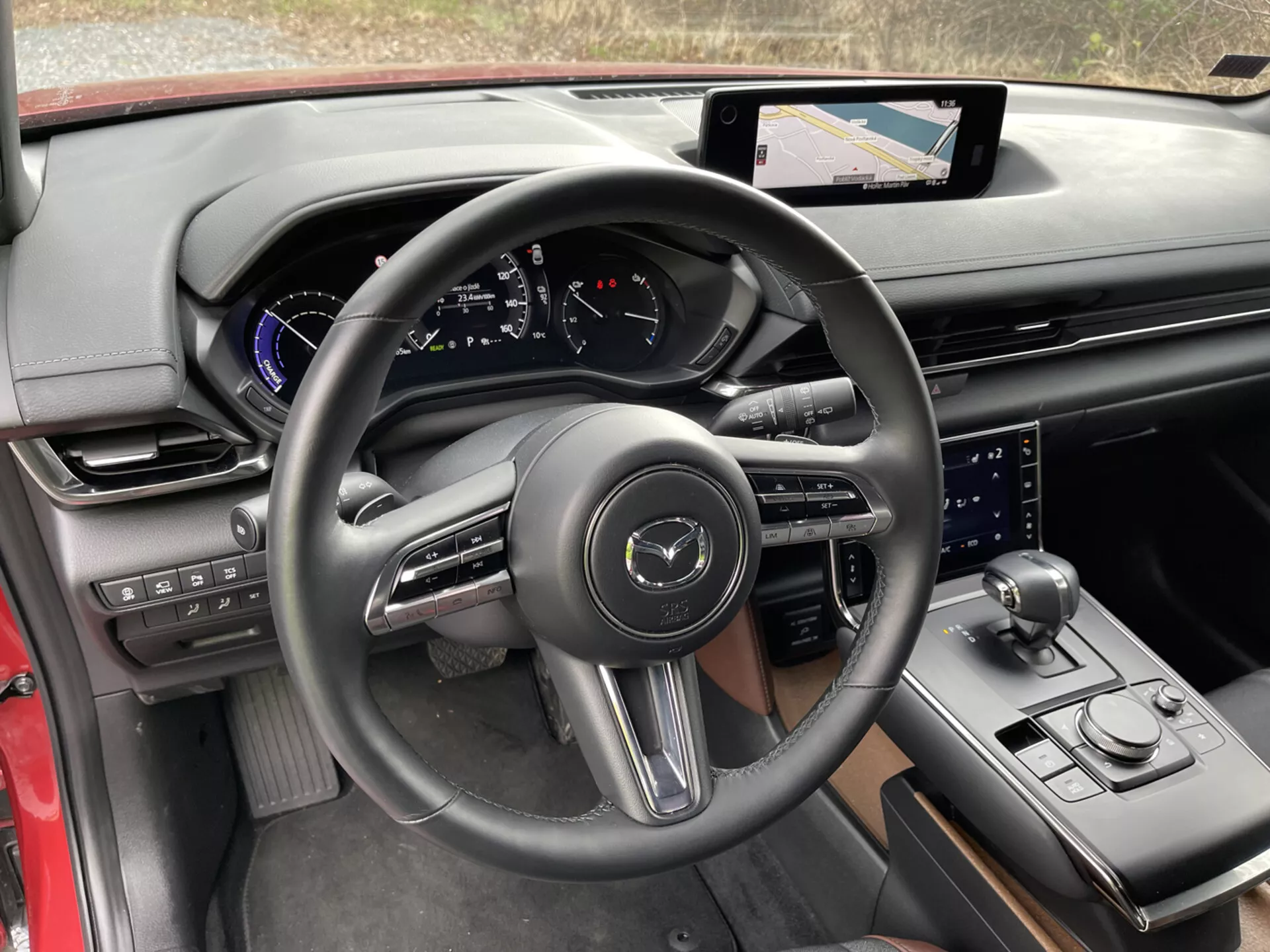Vista interior do Mazda MX-30 2020 a partir da perspectiva do motorista, focando em detalhes como o volante de três raios envolvido em couro com botões de controle e paddle shifts, cluster de instrumentos digital no painel com acentos azuis, sistema de infotainment com tela touch e navegação no console central, alavanca de câmbio esportiva, seleção de modo de condução, controle adicional para o sistema de infotainment, mistura luxuosa de materiais com acabamento preto e vermelho, porta-copos, e controles na porta para espelhos laterais e janelas, destacando a tecnologia moderna e design ergonômico.