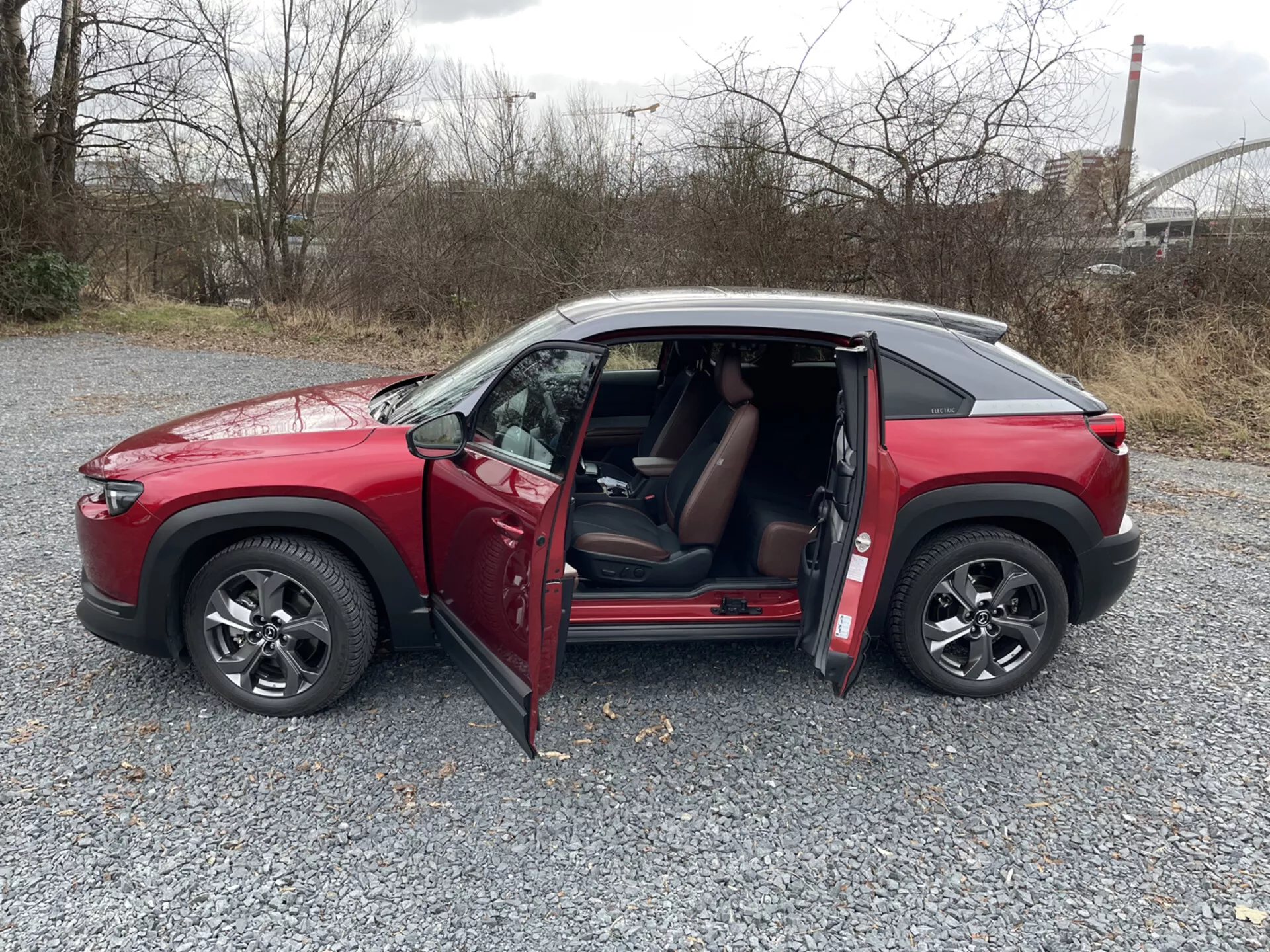 SUV Mazda MX-30 vermelho de quatro portas de 2020 em perfil lateral sobre cascalho com portas abertas, rodas de liga leve de múltiplos raios, janelas com película e teto de cor contrastante contra um fundo industrial e de vegetação.
